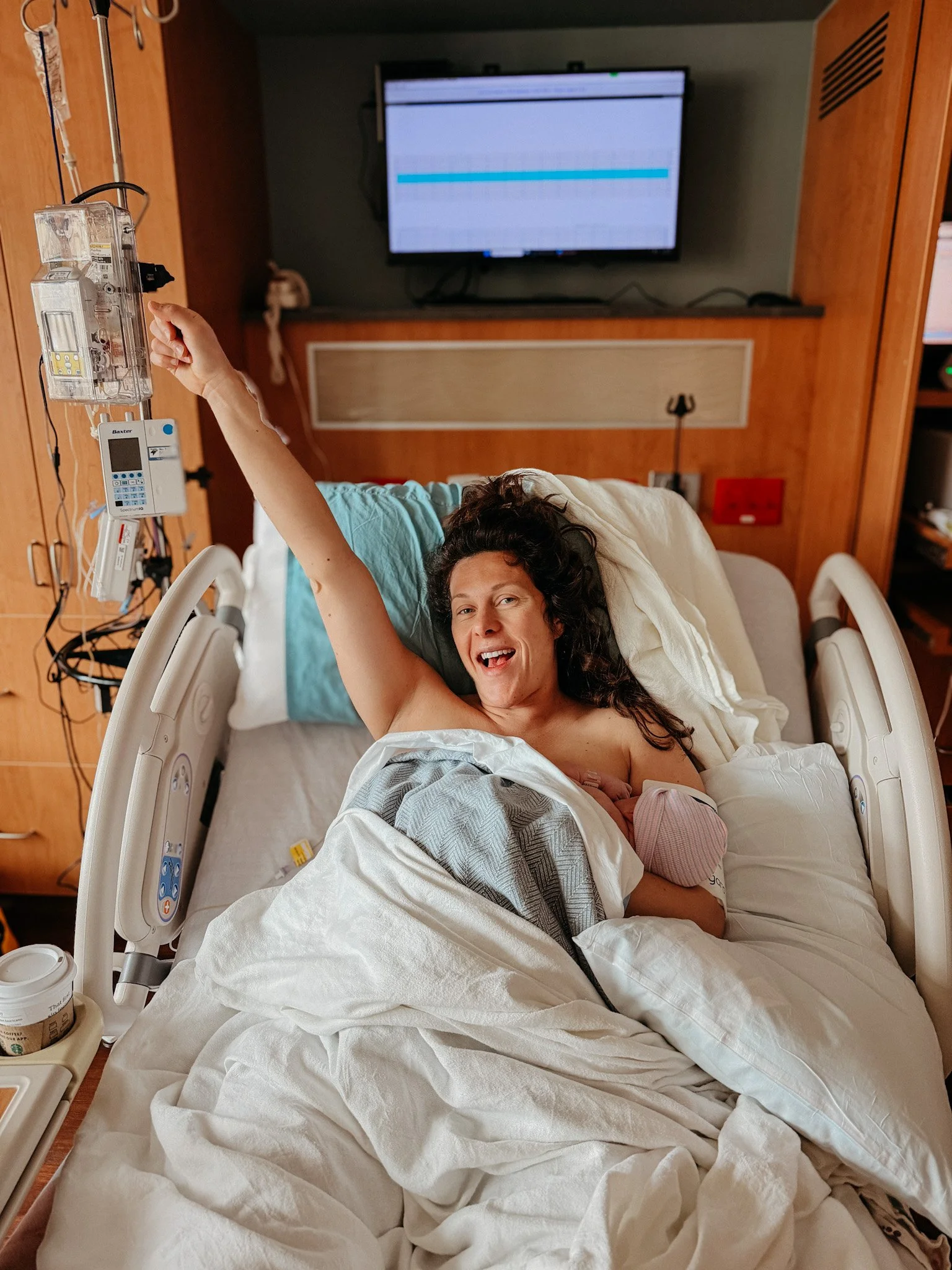 A woman lying in a hospital bed, smiling and reaching towards a monitor, with medical equipment and supplies around her.