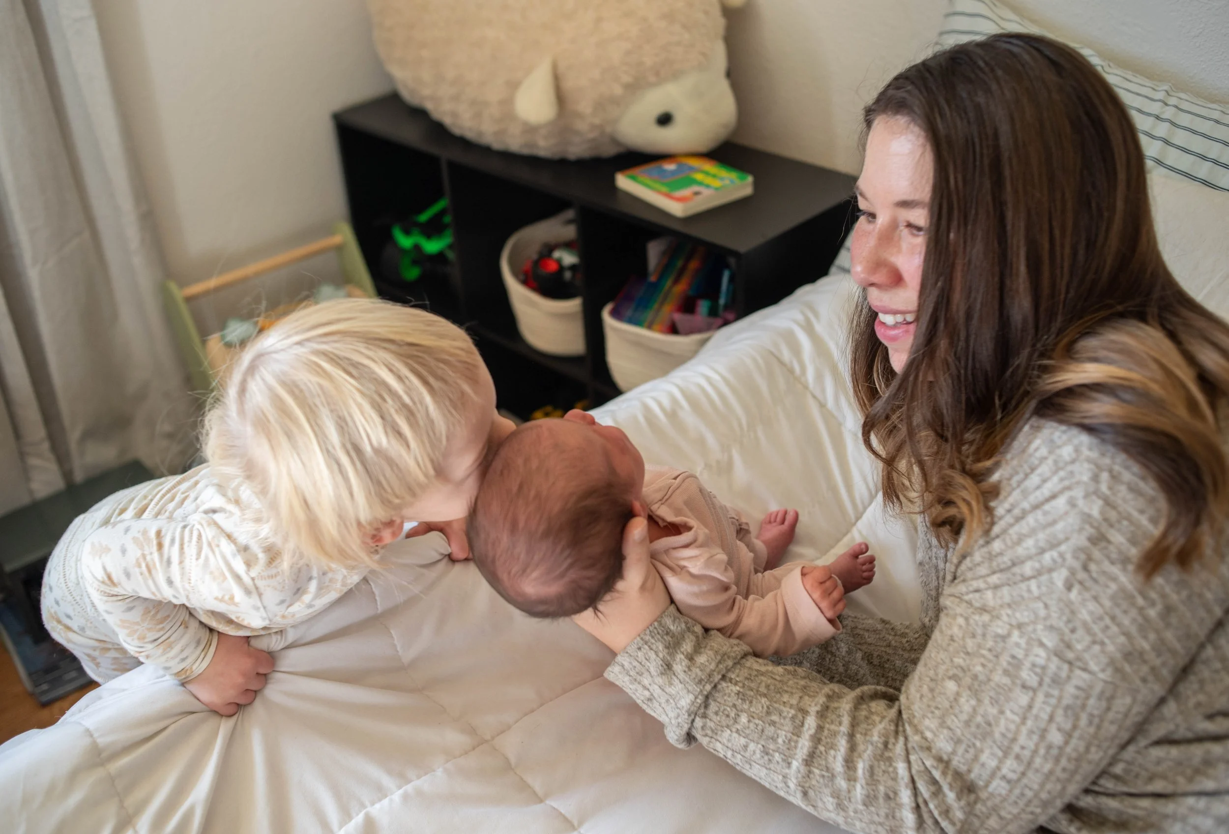 A woman and two children are on a bed, holding a baby. The woman is smiling and looking at the baby, who is lying in her arms. One child has blonde hair and is leaning over to kiss the baby, while the other child with dark hair is also involved. In the background, there is a black shelf with a large plush toy, books, and baskets.