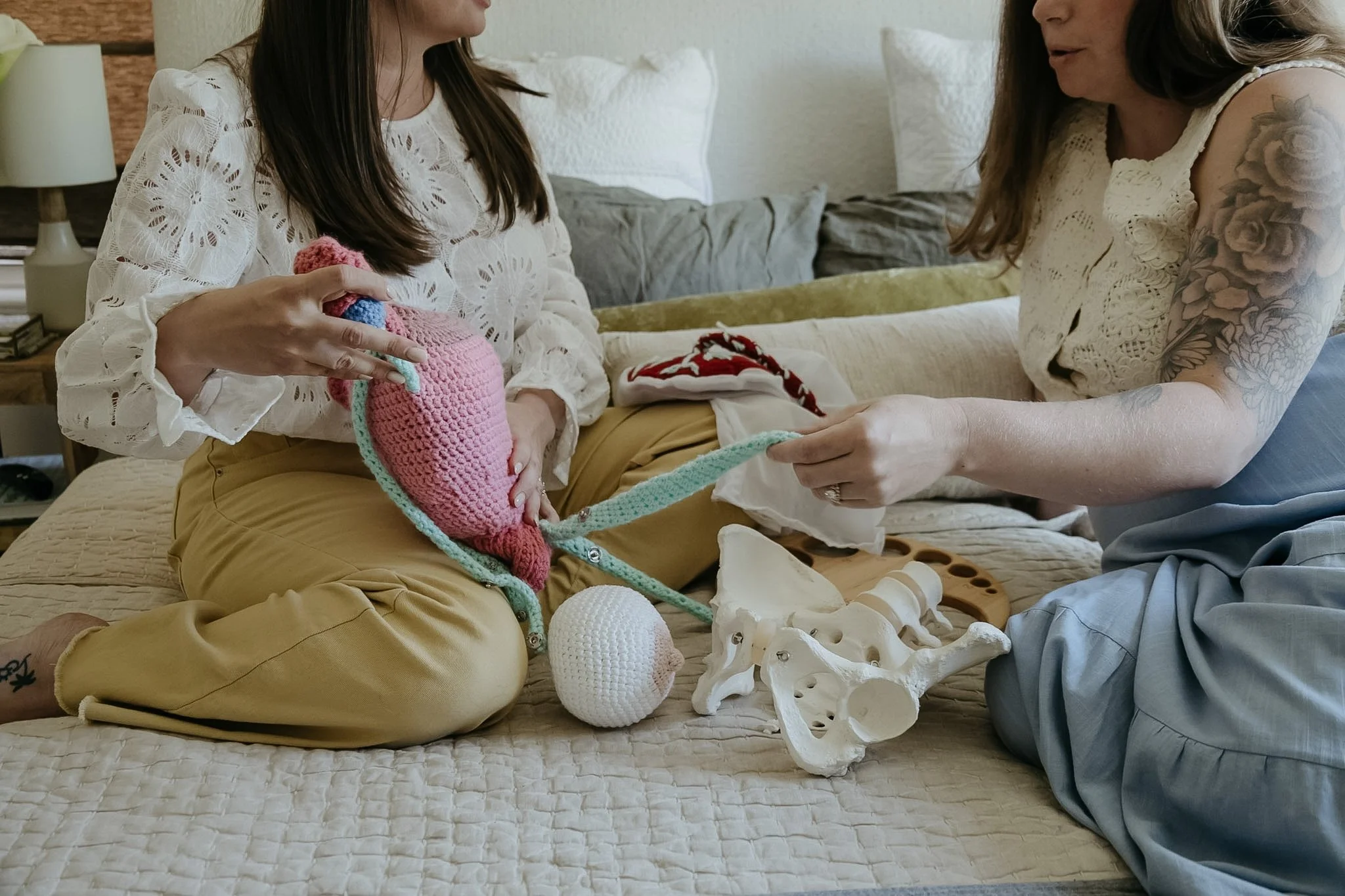 Two women sitting on a bed having a conversation surrounded by bones, skull, and crochet items, with one woman holding a pink and blue crocheted item.