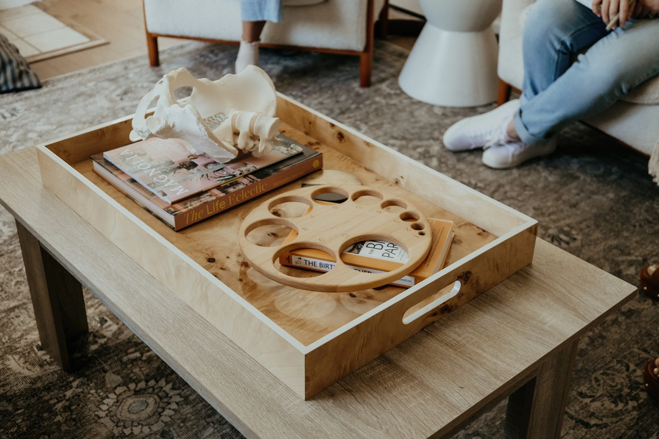 A wooden tray on a light-colored wooden table holds a skull model, a stack of magazines, and a wooden circular organizer with multiple holes. In the background, part of a person sitting on a sofa and a person sitting on the armrest wearing jeans and white sneakers are visible.