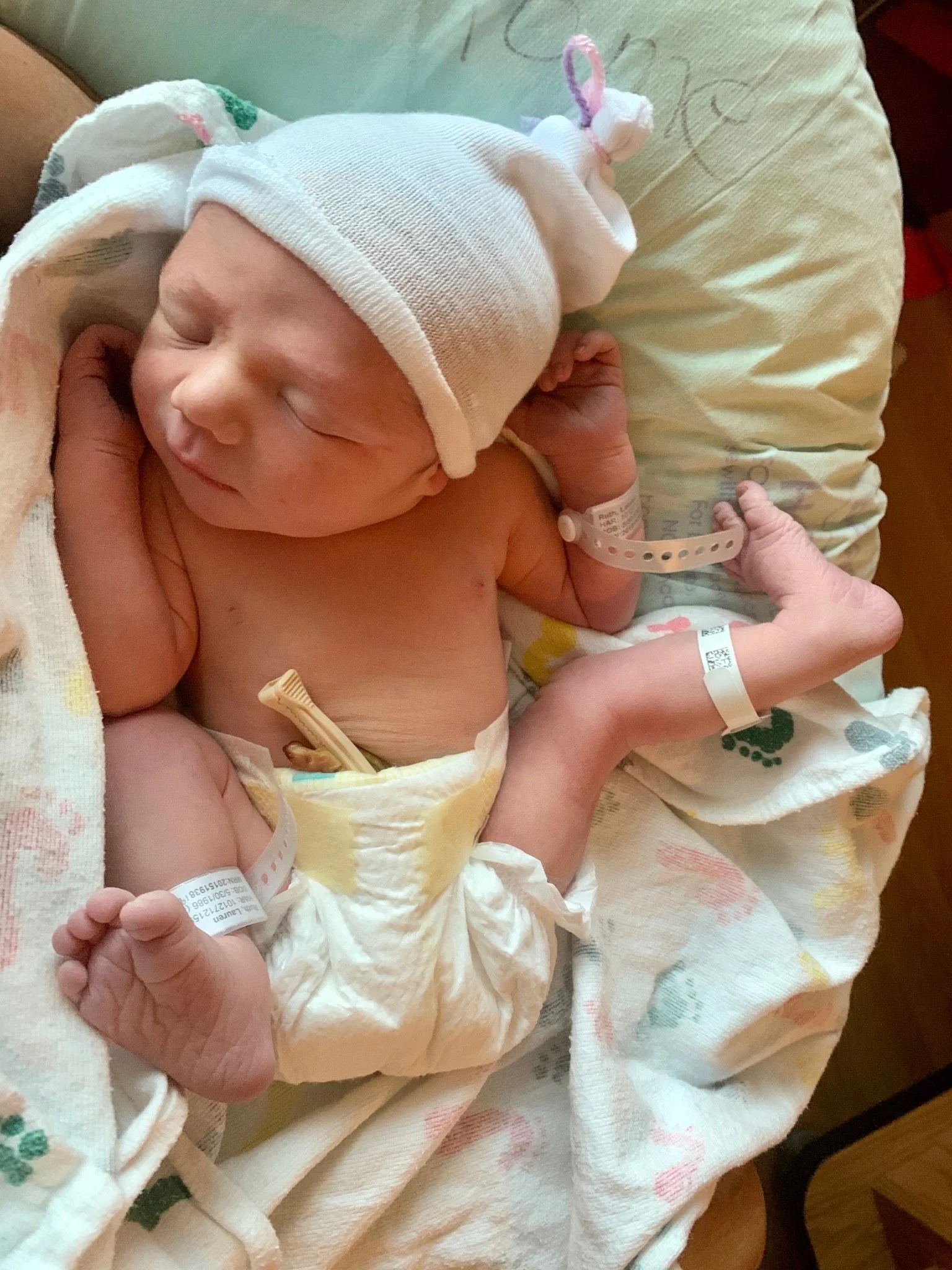 A newborn baby peacefully sleeping, wearing a white hat with a pink and purple ribbon, and wrapped in a hospital blanket. The baby has a hospital bracelet on their wrist and is holding a small object with a label.