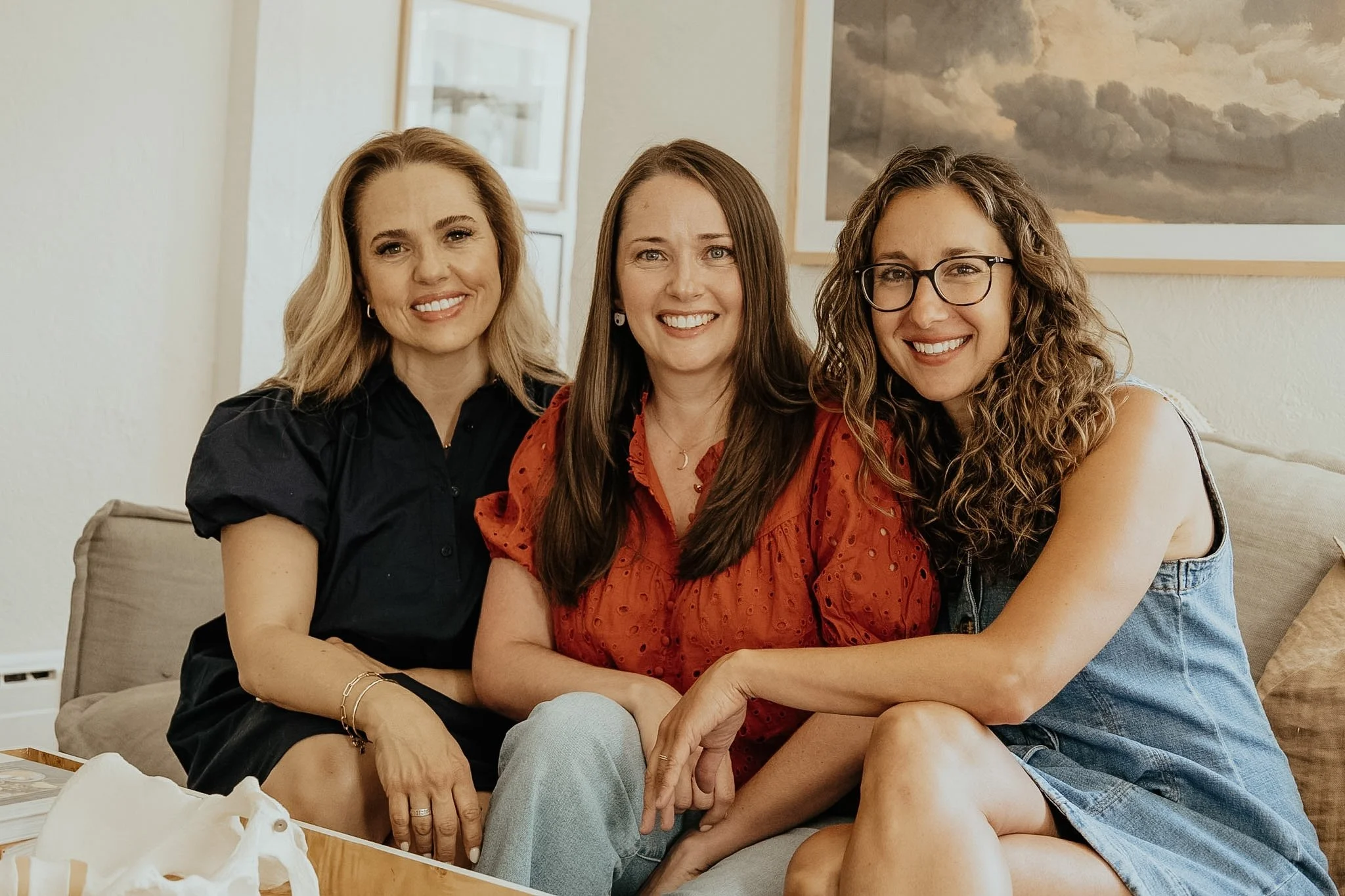 Three women sitting closely together on a beige couch, smiling at the camera. The woman on the left has blond hair and is wearing a black top. The woman in the middle has long brown hair and is wearing a red blouse. The woman on the right has curly brown hair, glasses, and is wearing a sleeveless top with denim shorts. There is a painting of a cloudy sky on the wall behind them and some tissue papers on a wooden tray in the foreground.