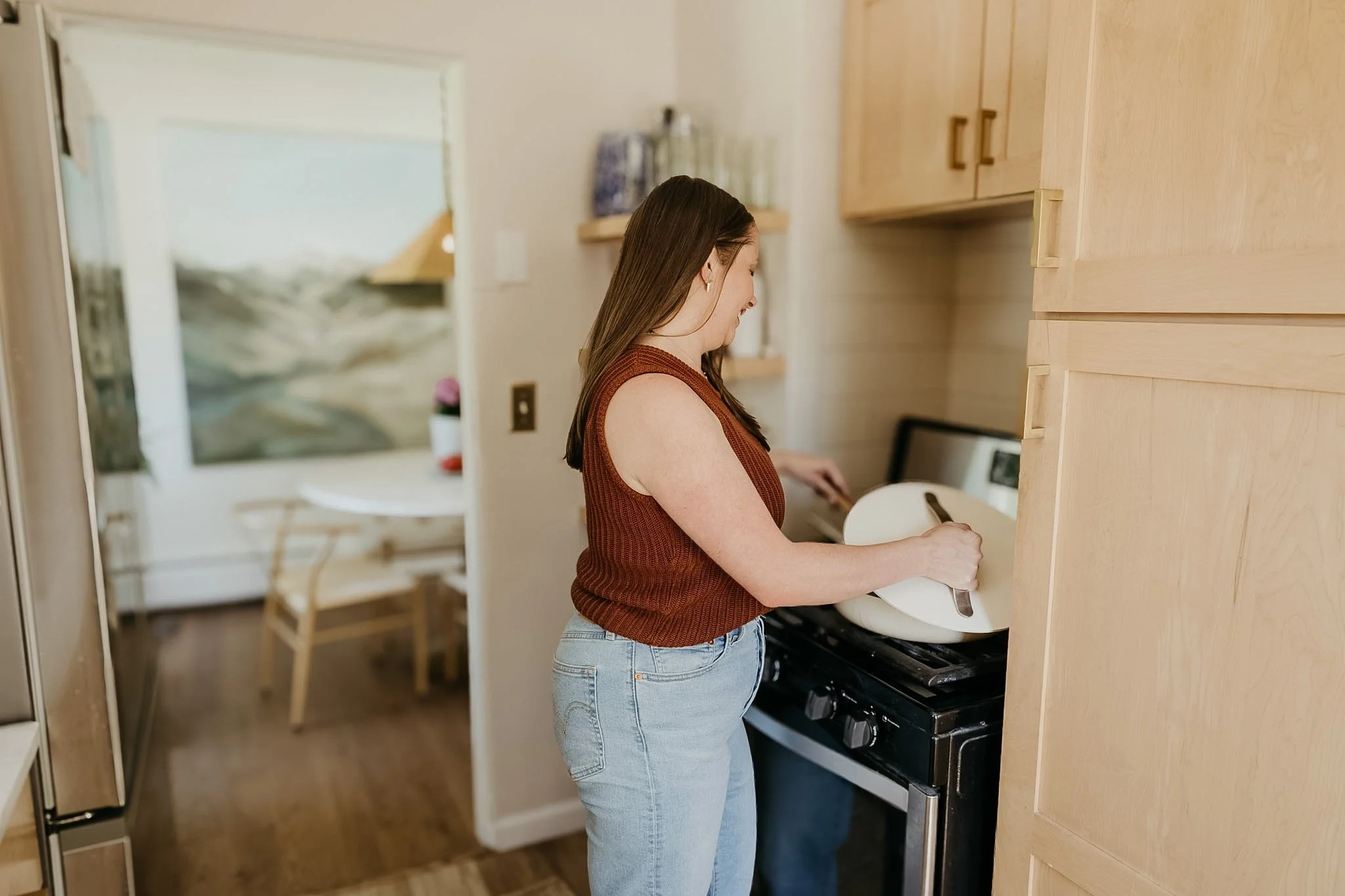 A woman with long brown hair wearing a sleeveless rust-colored top and blue jeans is standing at a stove, toasting bread using a toaster in a kitchen with wooden cabinets.