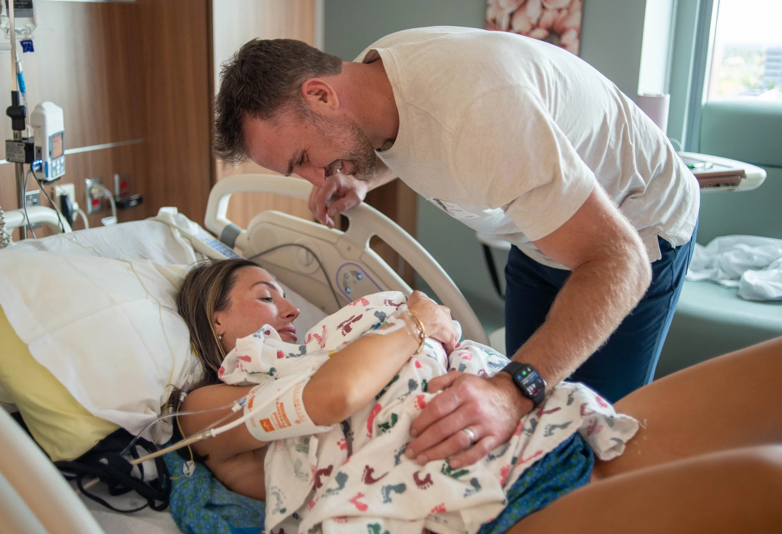A man with short hair and a beard leaning over a hospital bed, smiling and holding hands with a woman lying in the bed. The woman is wearing hospital gown and a wristband, with a breathing tube connected to her arm. Medical equipment is visible in the background, and sunlight is coming through a window.