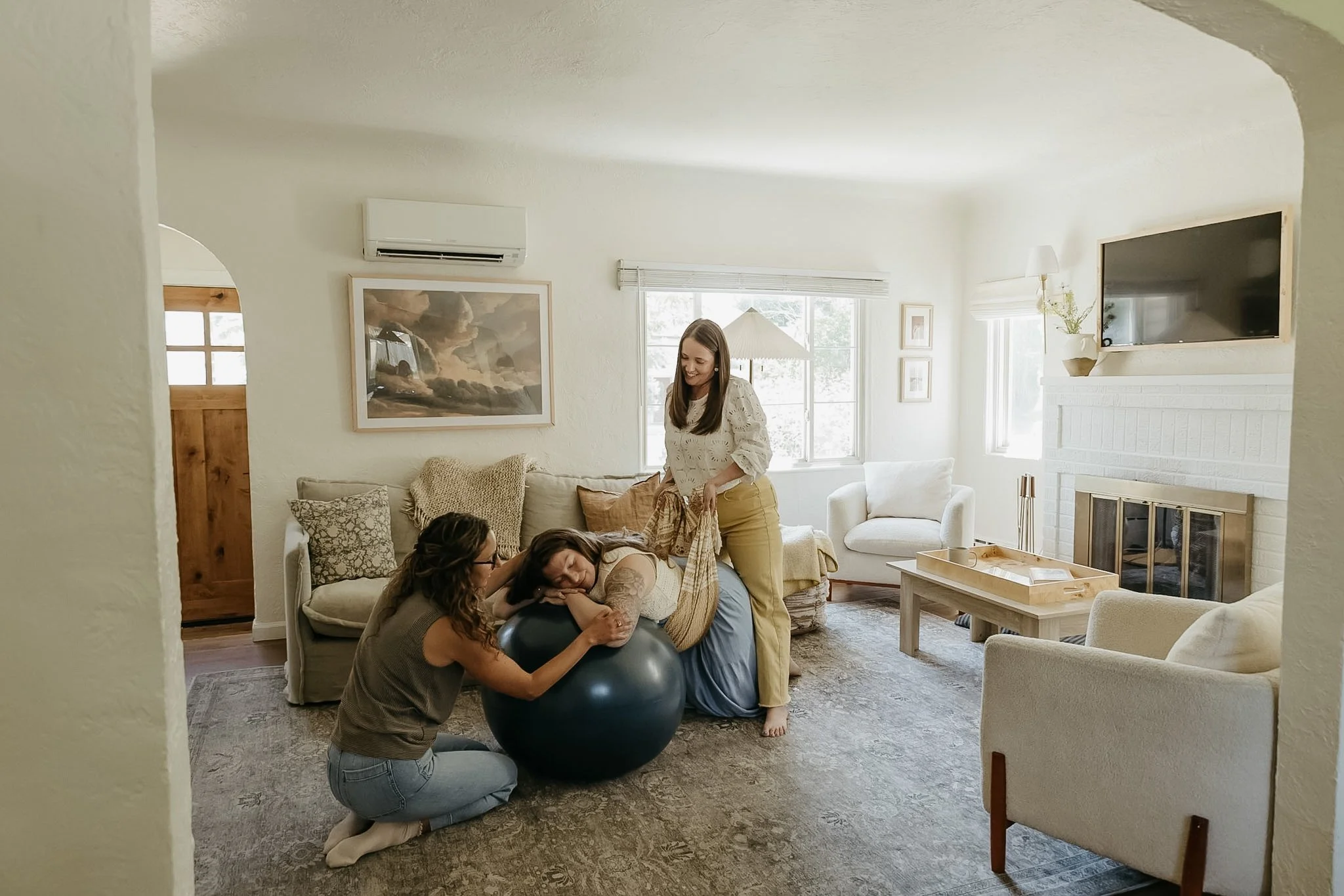 A woman in a white blouse and yellow pants is standing in a living room, smiling as she interacts with three women who are sitting on a couch. One woman is kneeling next to a yoga ball, and two others are lying on the couch, one with dark curly hair and the other with glasses and long hair. The living room has beige walls, a white fireplace, a wall-mounted TV, and large windows with blinds.