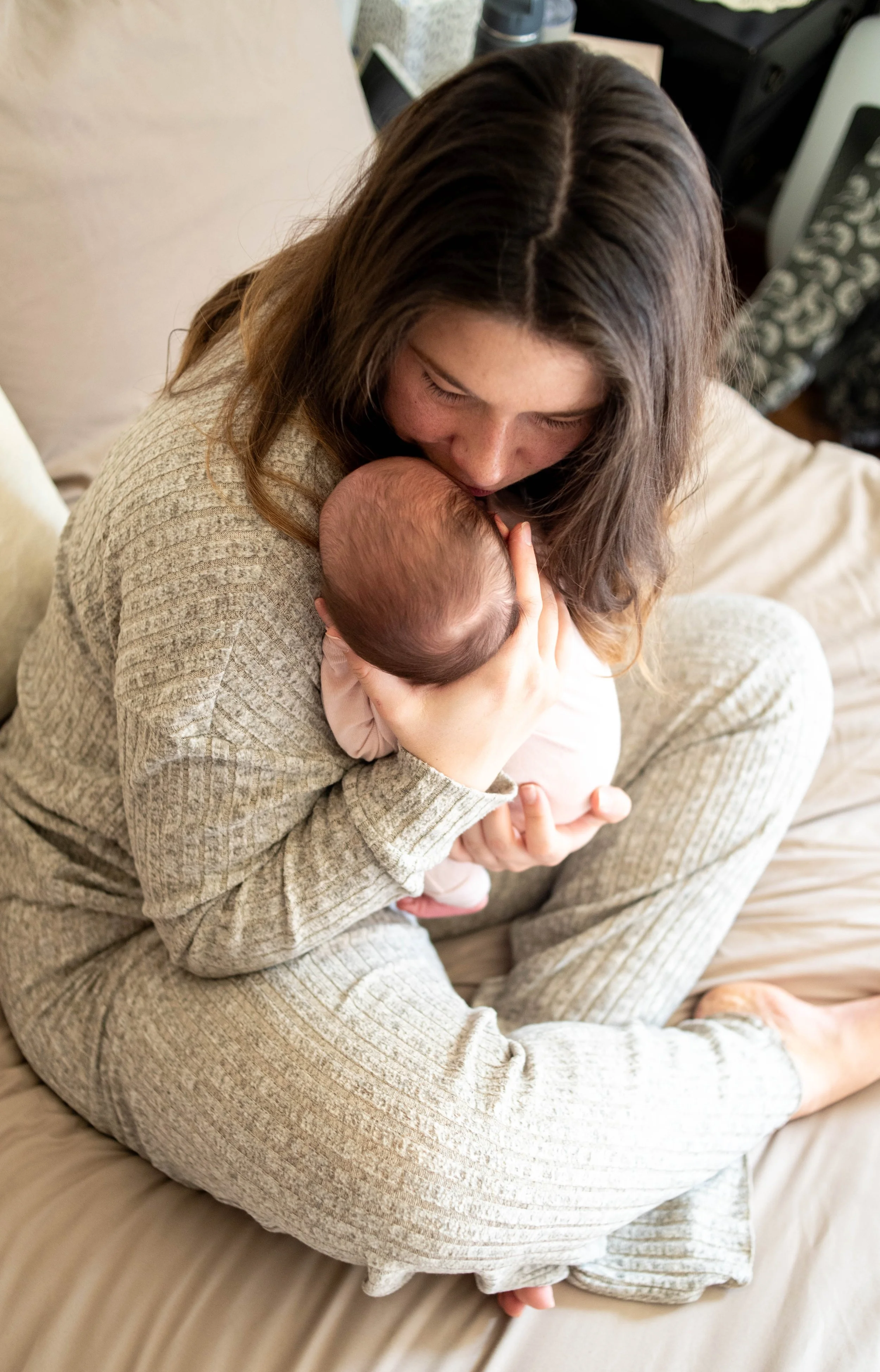 A woman with long brown hair sitting on a bed, holding a baby close to her chest, gently kissing the baby's head in a cozy bedroom setting.