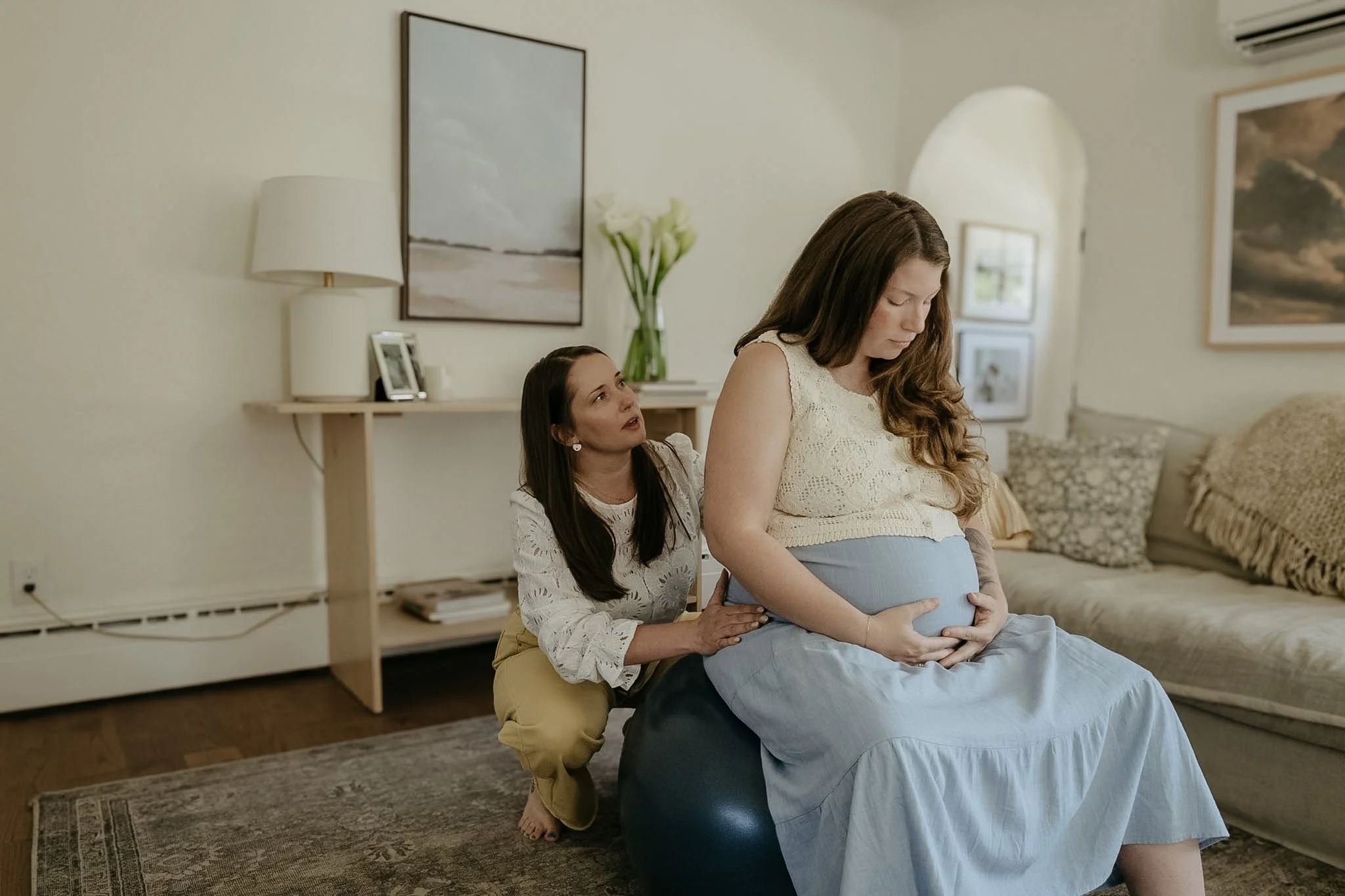 A woman sits on a stool, cradling her pregnant belly, while another woman kneels behind her with her hands on her shoulders in a cozy living room.