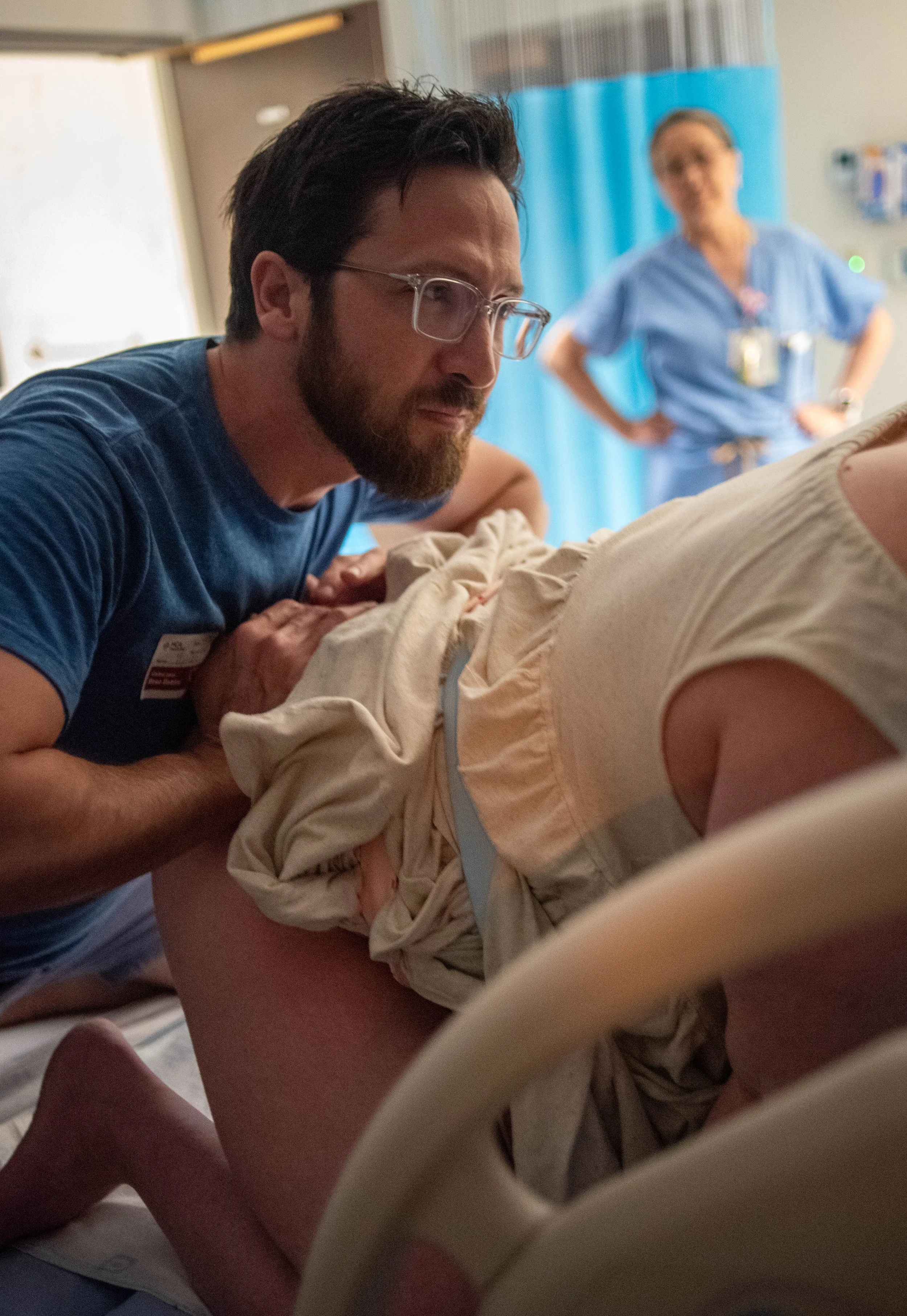 Medical professional performing a procedure on a patient in a hospital room with a nurse observing in the background.