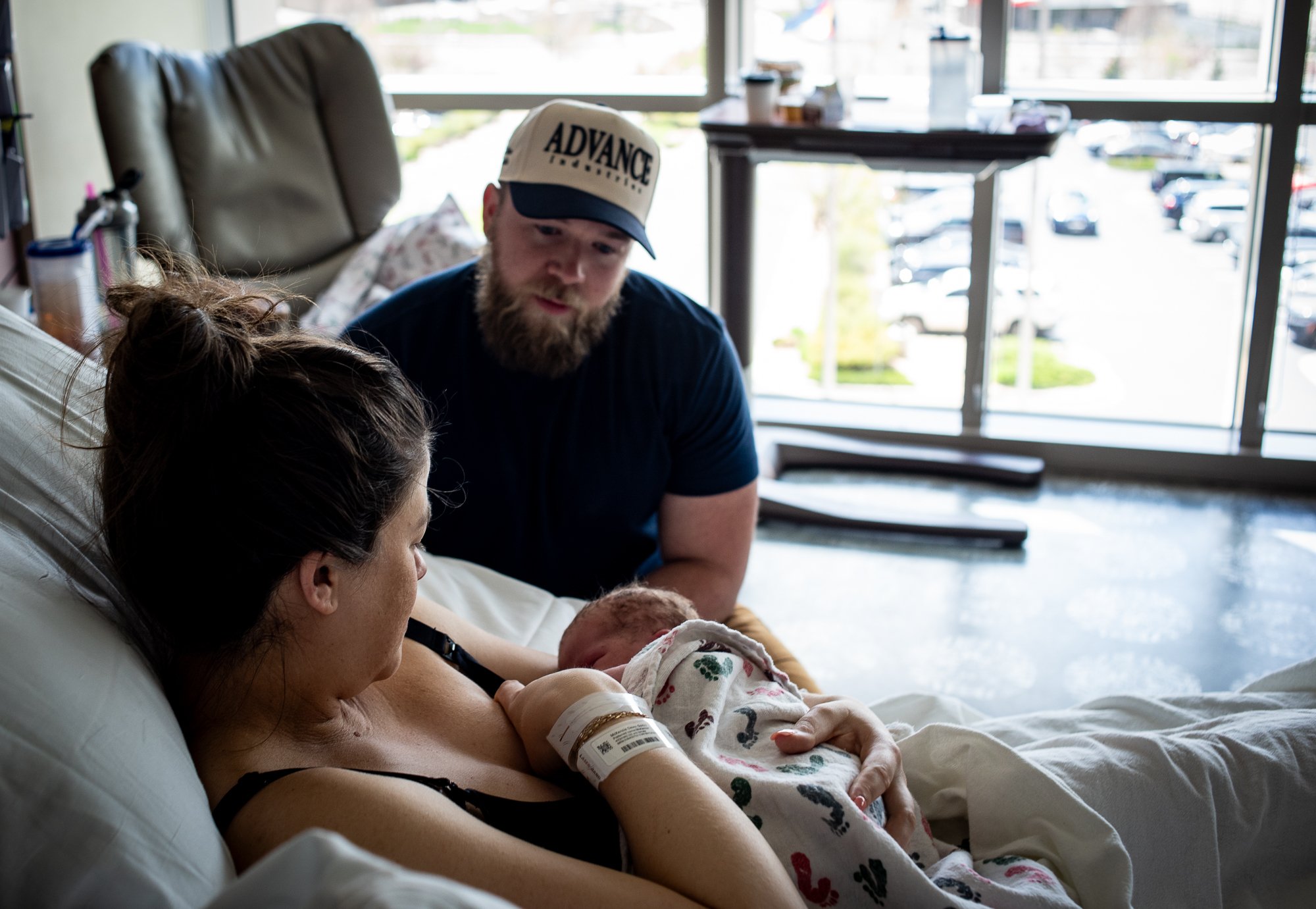 A woman lying in a hospital bed holding a newborn baby, with a man in a cap looking on in a hospital room with large windows and parking lot outside.