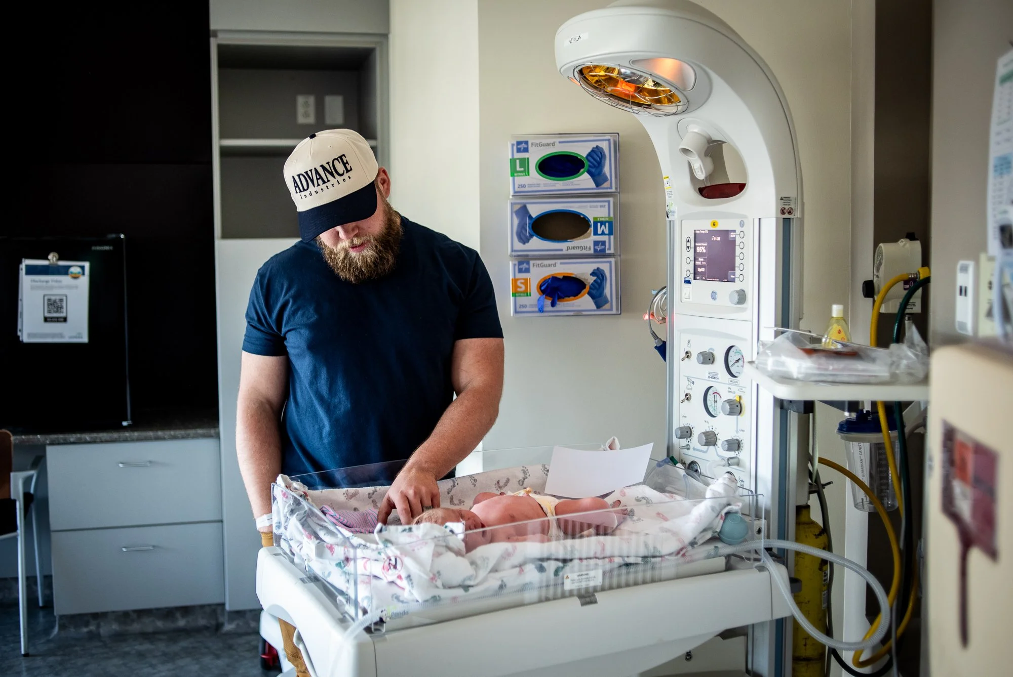 A man with a beard wearing a white cap and navy T-shirt is standing next to a hospital bassinet with a newborn baby inside. The man appears to be attending to the baby in a hospital room equipped with medical devices and supplies.
