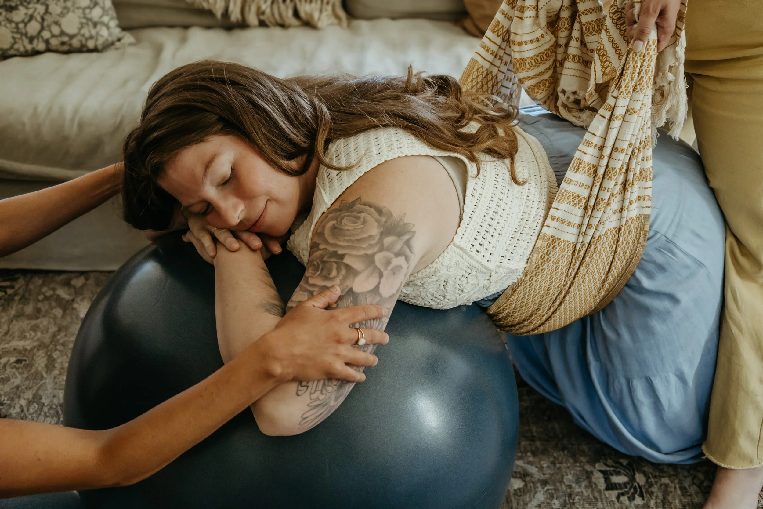 Woman with tattoos receiving a tattoo on her arm while lying on a massage table in a cozy room.