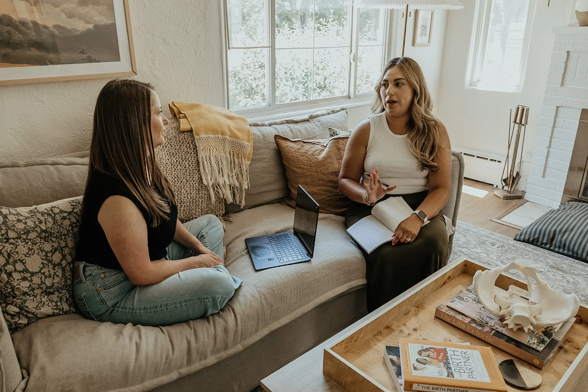 Two women sitting on a beige couch having a conversation, with a window behind them and a wooden table in front of them holding books and a skull model.