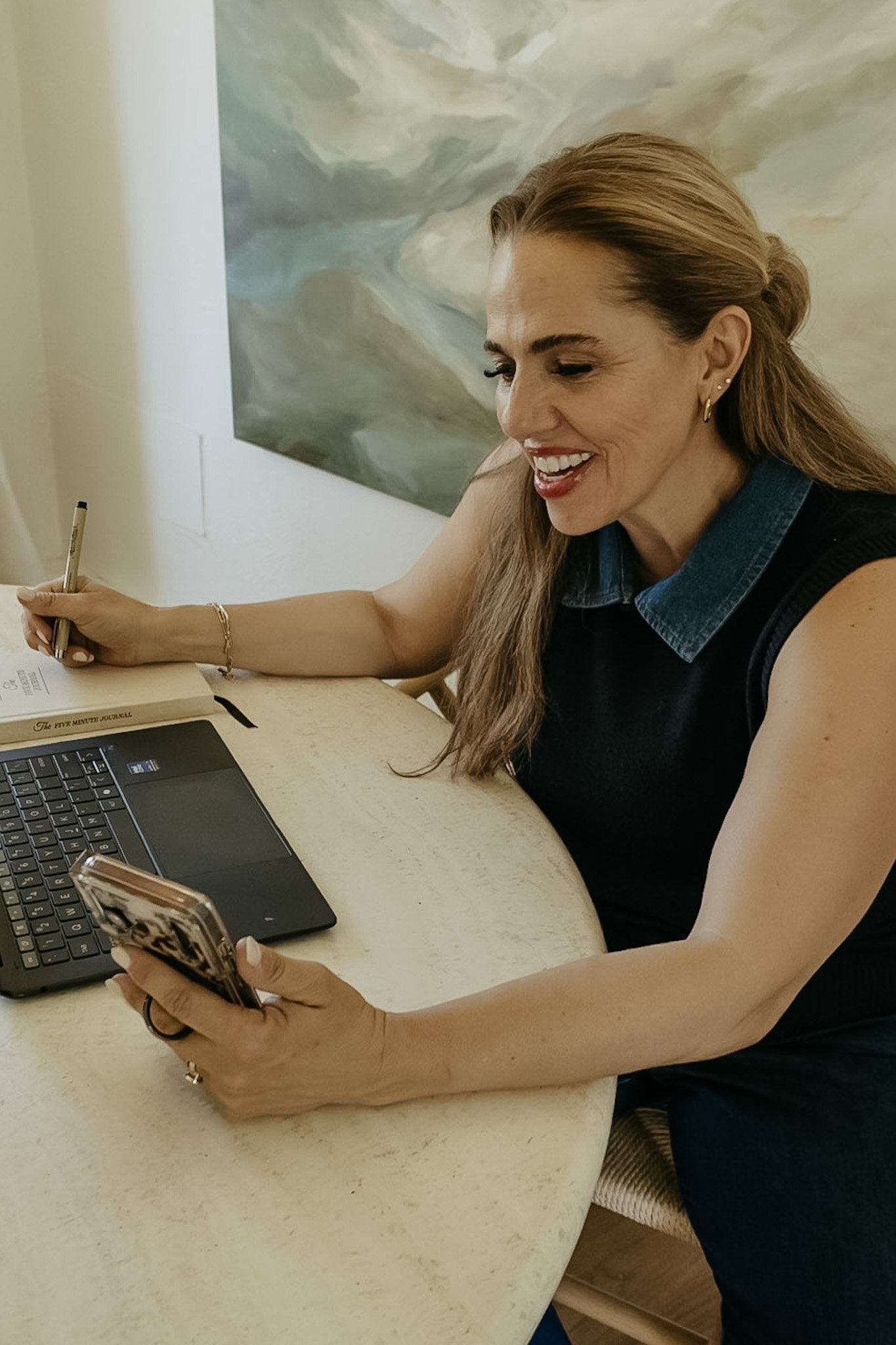 A woman with long red hair, wearing a sleeveless black top and denim collar, is smiling and writing in a notebook at a table. She is also holding a smartphone, with a laptop and a painting of an abstract scene in the background.