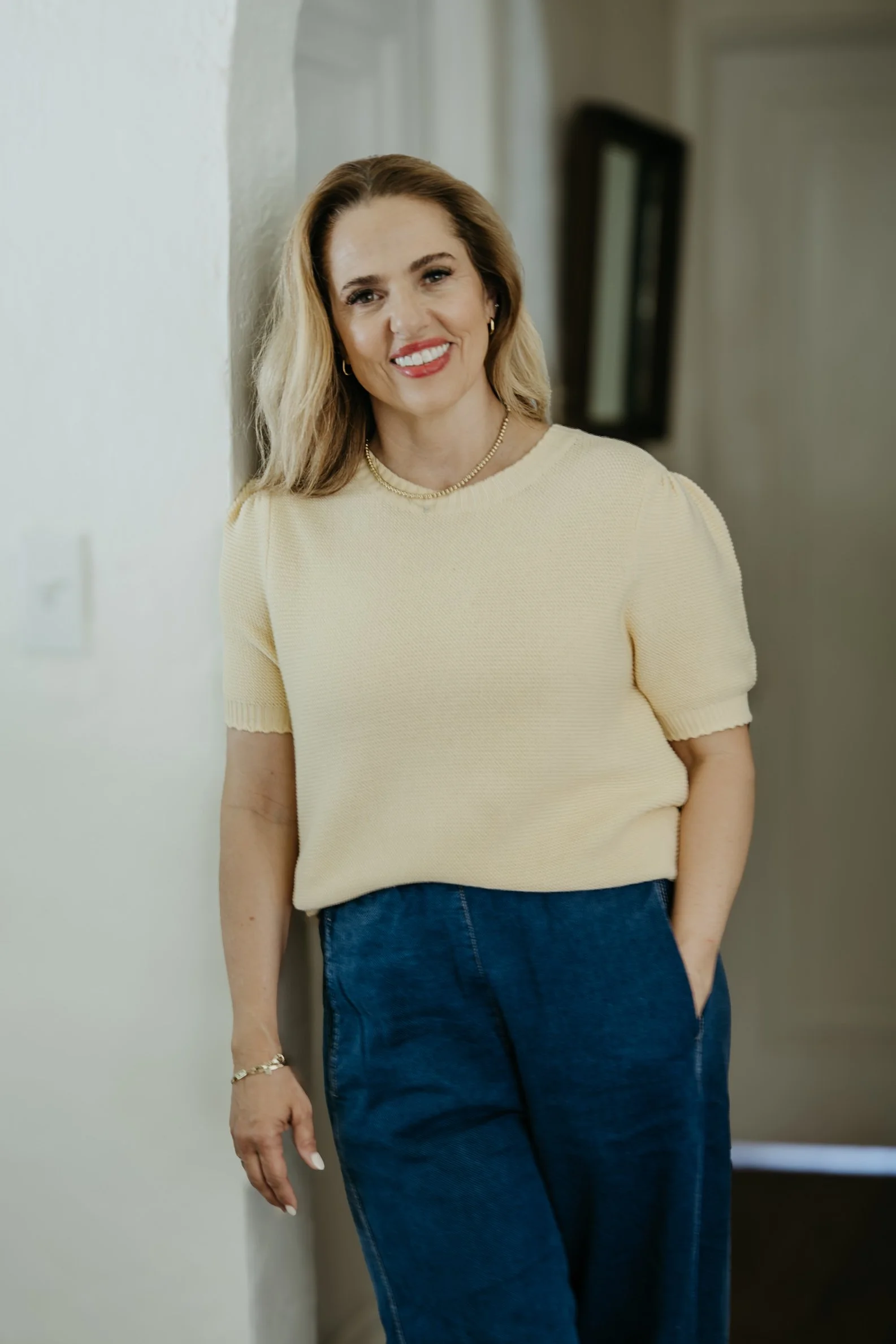 A woman with blonde hair and a bright smile, wearing a cream-colored short-sleeve sweater and blue jeans, standing against a white wall inside a house.