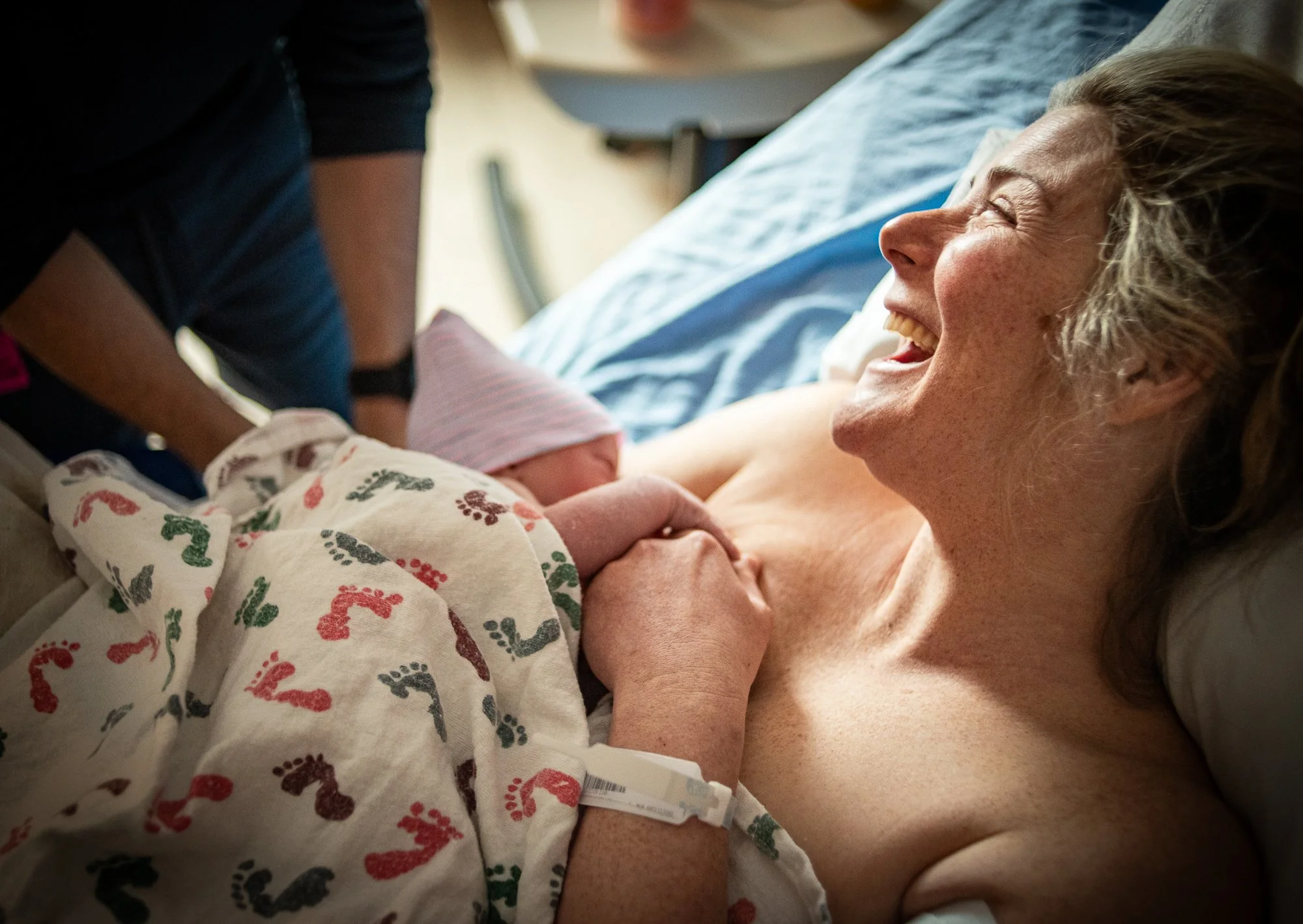 A woman in labor holding her newborn baby in a hospital bed, with a person assisting beside her.