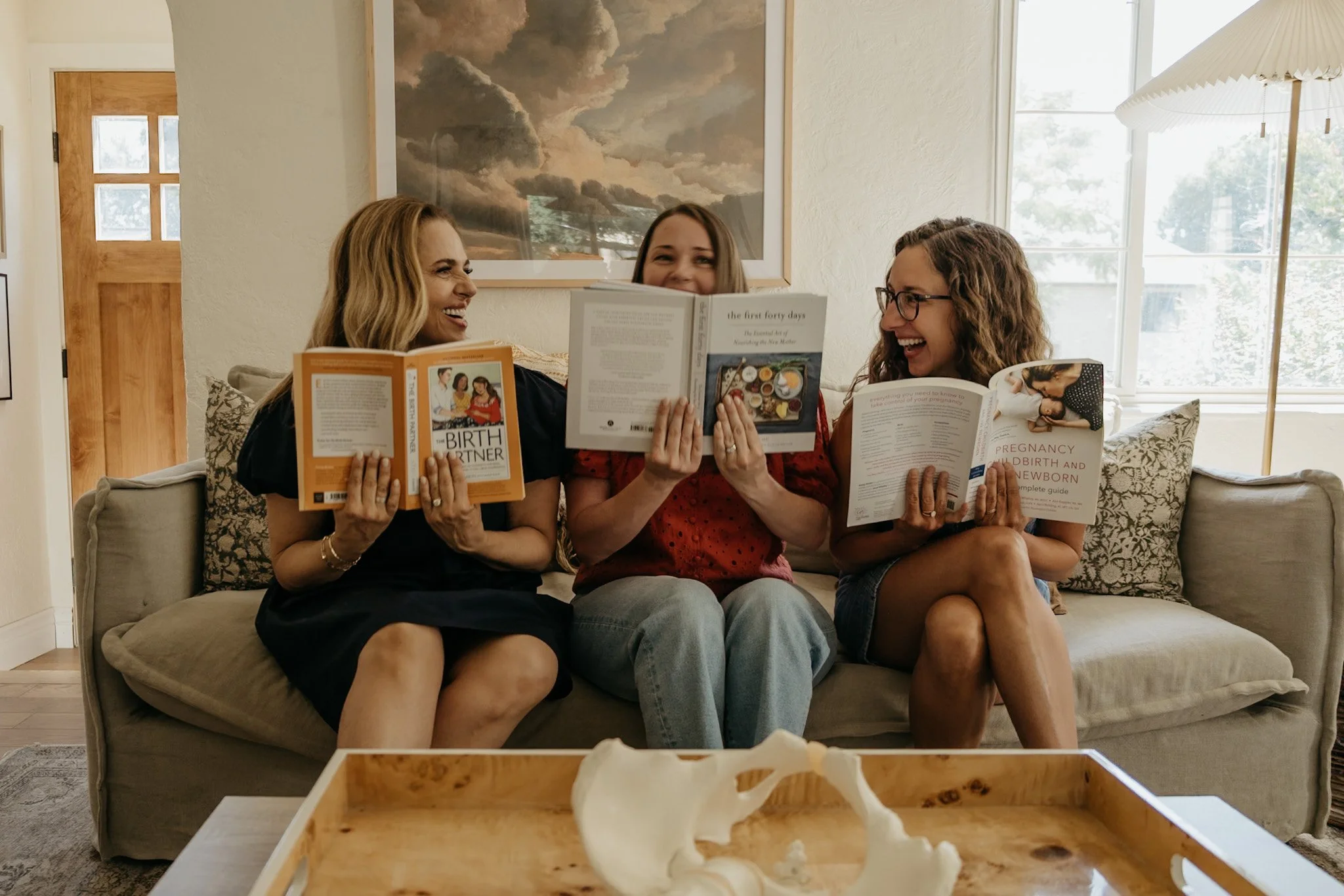 Three women sitting on a beige couch in a bright living room, reading pregnancy and childbirth books, laughing and smiling together.