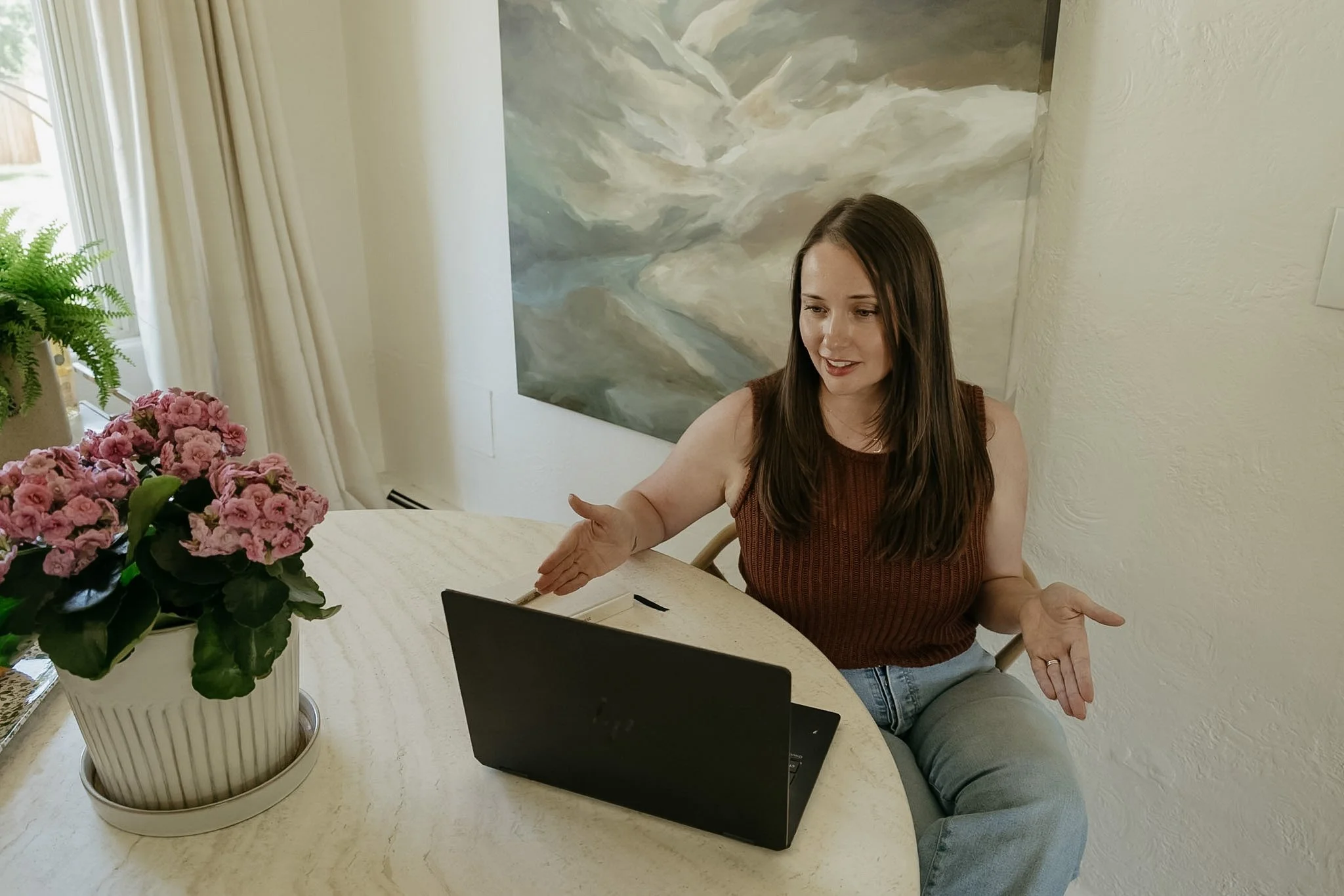 A woman with long brown hair, wearing a sleeveless brown top and jeans, is sitting at a table with a laptop in front of her. She is gesturing with her hands while speaking or explaining something. There is a large potted pink flower and a green fern on the table. Behind her is a wall with a cloudy landscape painting and a window with white curtains, allowing natural light into the room.