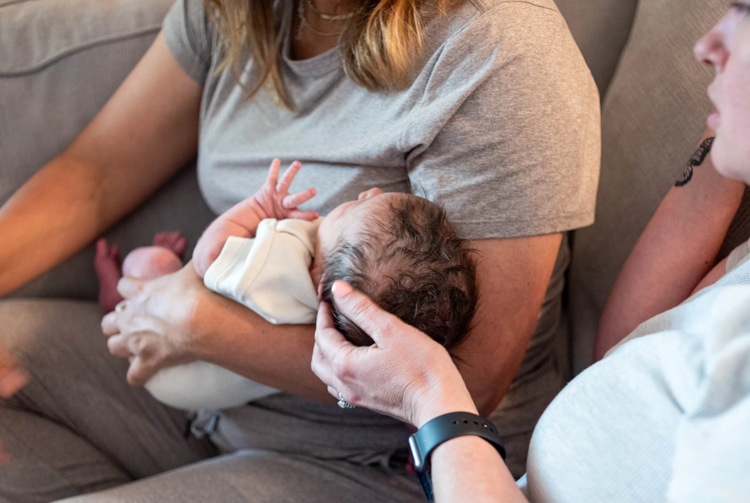 A woman holding a newborn baby on her lap, with another woman beside her gently supporting the baby's head, on a brown couch.