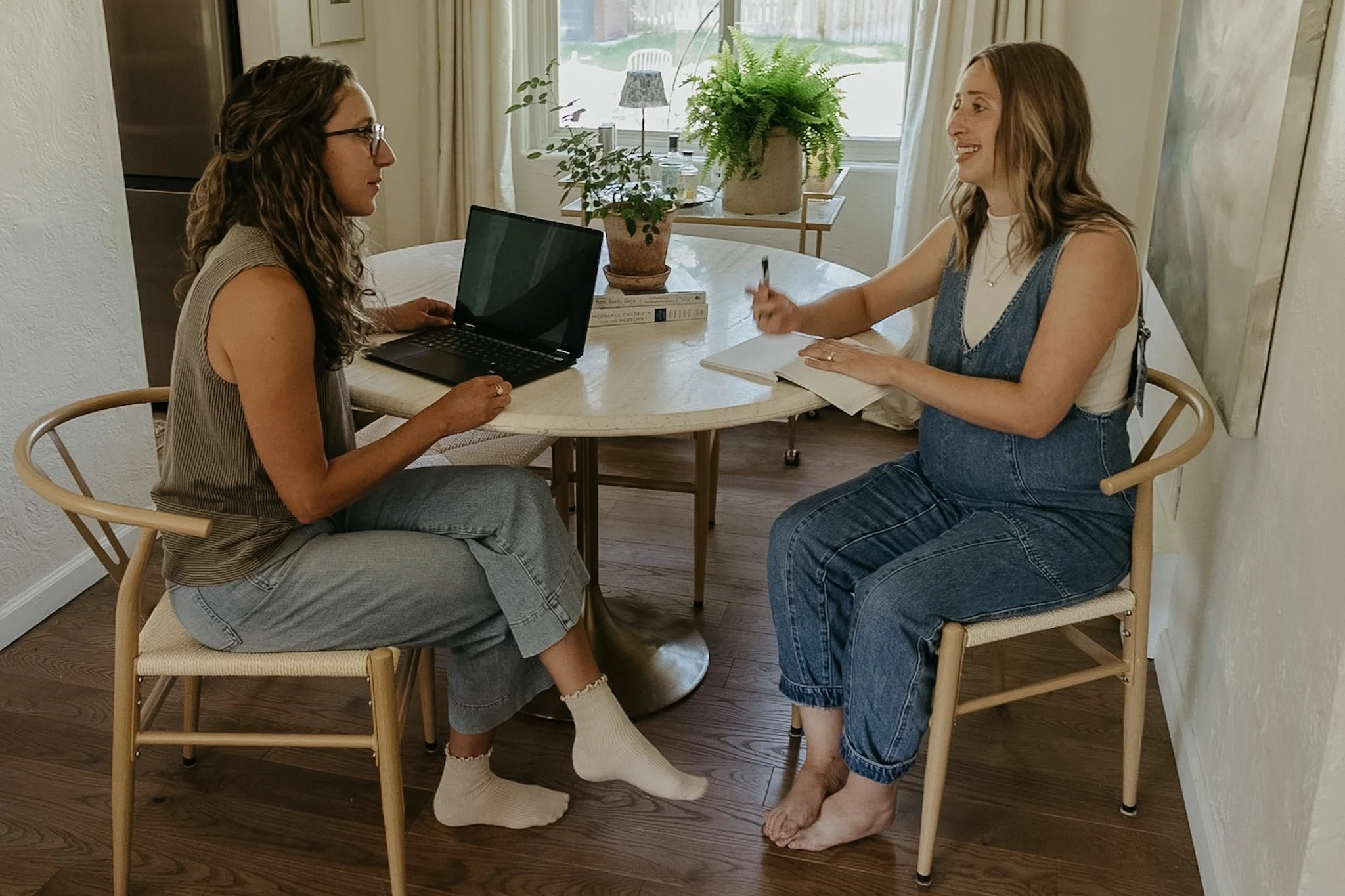 Two women sitting at a small round table in a cozy, sunlit room. One woman is on the left with glasses and curly hair, looking at the other woman. The woman on the right has long hair, is smiling, and is gesturing with her right hand. There is a laptop, a potted fern, some books, and a notebook on the table. The background shows a window with curtains, a houseplant, and a standing lamp.