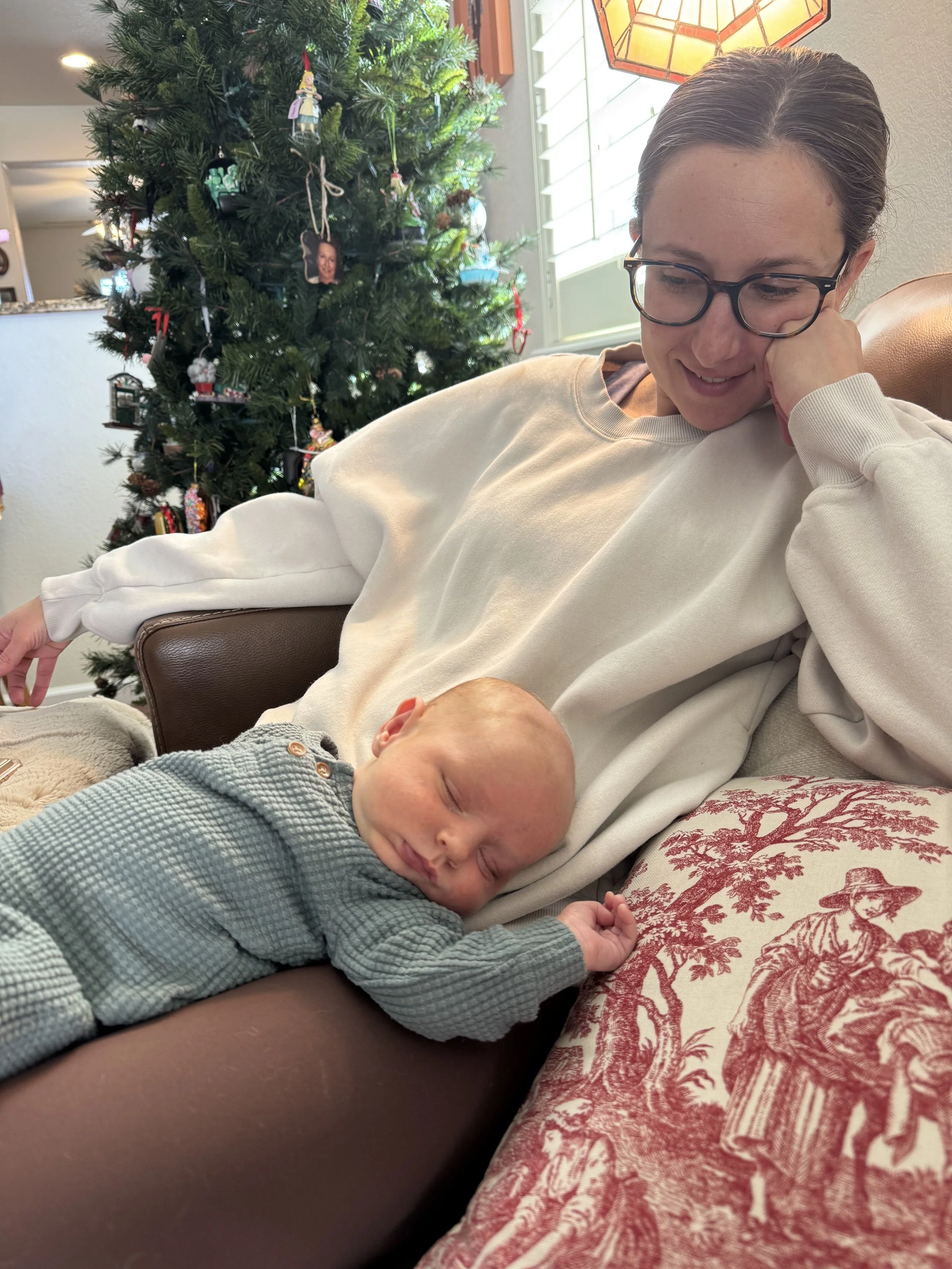 A woman with glasses and brown hair tied back, smiling gently, as she leans her head on her hand while sitting on a chair. A sleeping baby dressed in a gray and white checkered outfit is resting on her lap. In the background, a decorated Christmas tree with various ornaments and a lamp with a stained glass shade are visible, along with a window with white shutters.