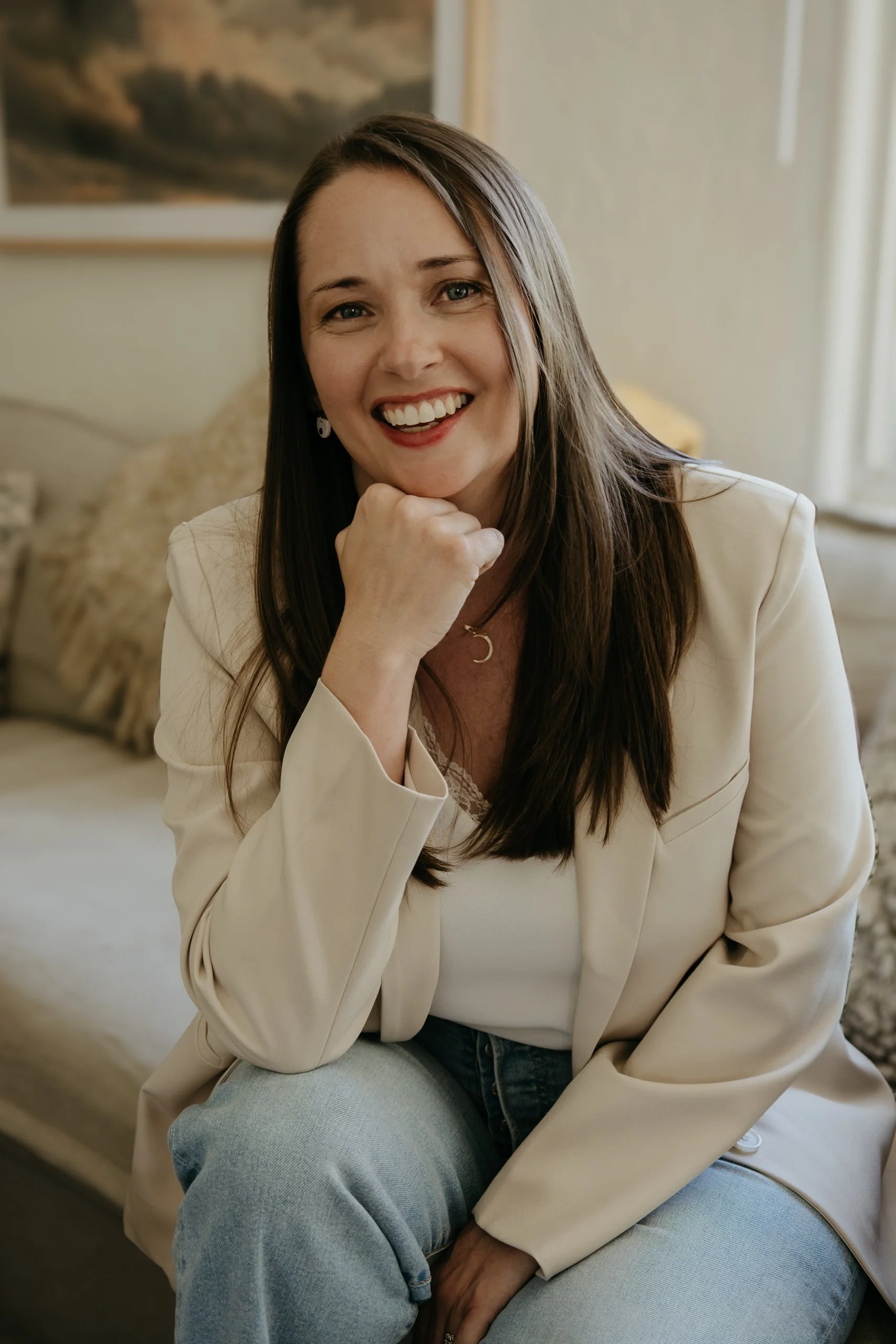 A woman with long brown hair smiling and resting her chin on her hand, sitting in a cozy room with neutral decor, wearing a cream blazer and light blue jeans.