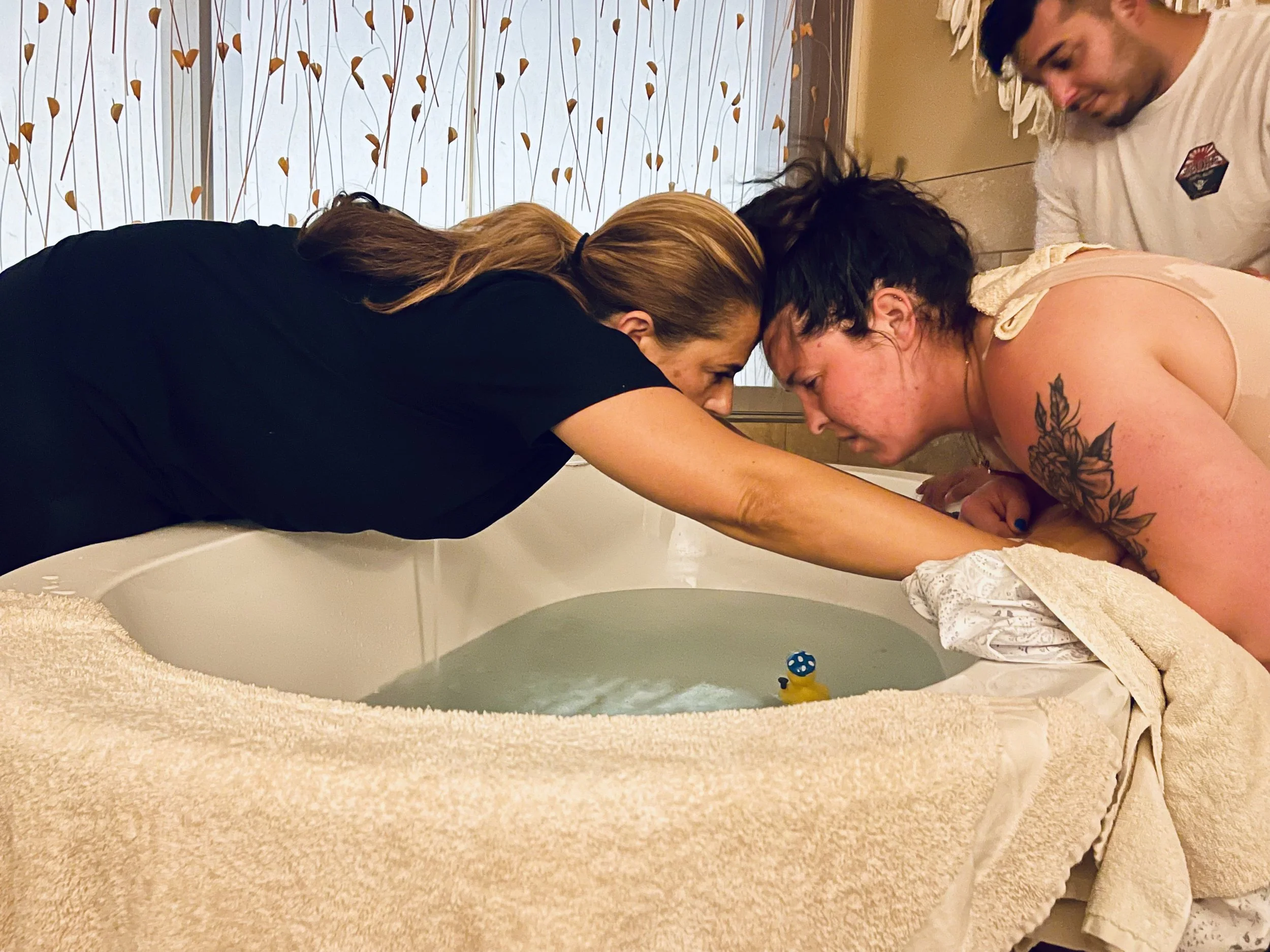 Two people are leaning over a bathtub, facing each other with foreheads touching, engaging in a close, intimate moment. A third person, smiling, observes from the right side of the image. The background features a wall with a floral pattern, and the foreground shows the edge of the bathtub with a towel and a yellow rubber duck.