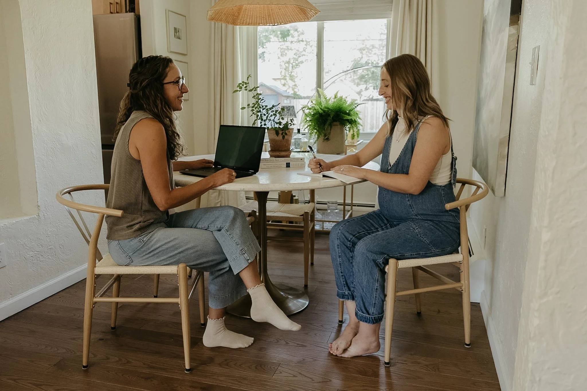 Two women are in a bright kitchen, sitting at a round table, smiling and engaging in conversation. One woman has curly brown hair, glasses, and is wearing a beige top and sweatpants. The other woman has long wavy hair, and is wearing overalls and a white tank top. There are green plants on the table, and a laptop in front of the woman on the left. The background features a window with curtains, a fridge, and some artwork on the walls.