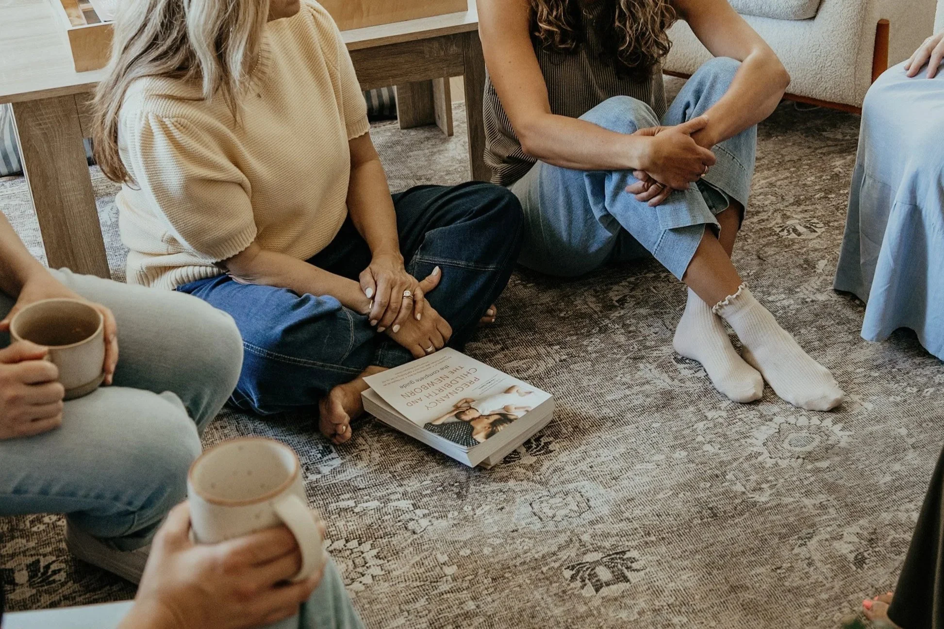 People sitting on a carpeted floor in a circle, holding cups, with a book titled 'Pregnancy and Childbirth in the Newborn Age' in front of them. The scene appears to be a group discussion or workshop.