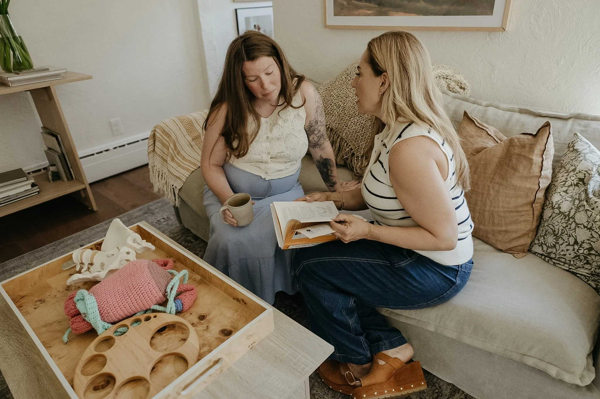 Two women sitting on a sofa in a cozy living room, one showing a book to the other, with a wooden tray of handmade knitted items on a table in front.