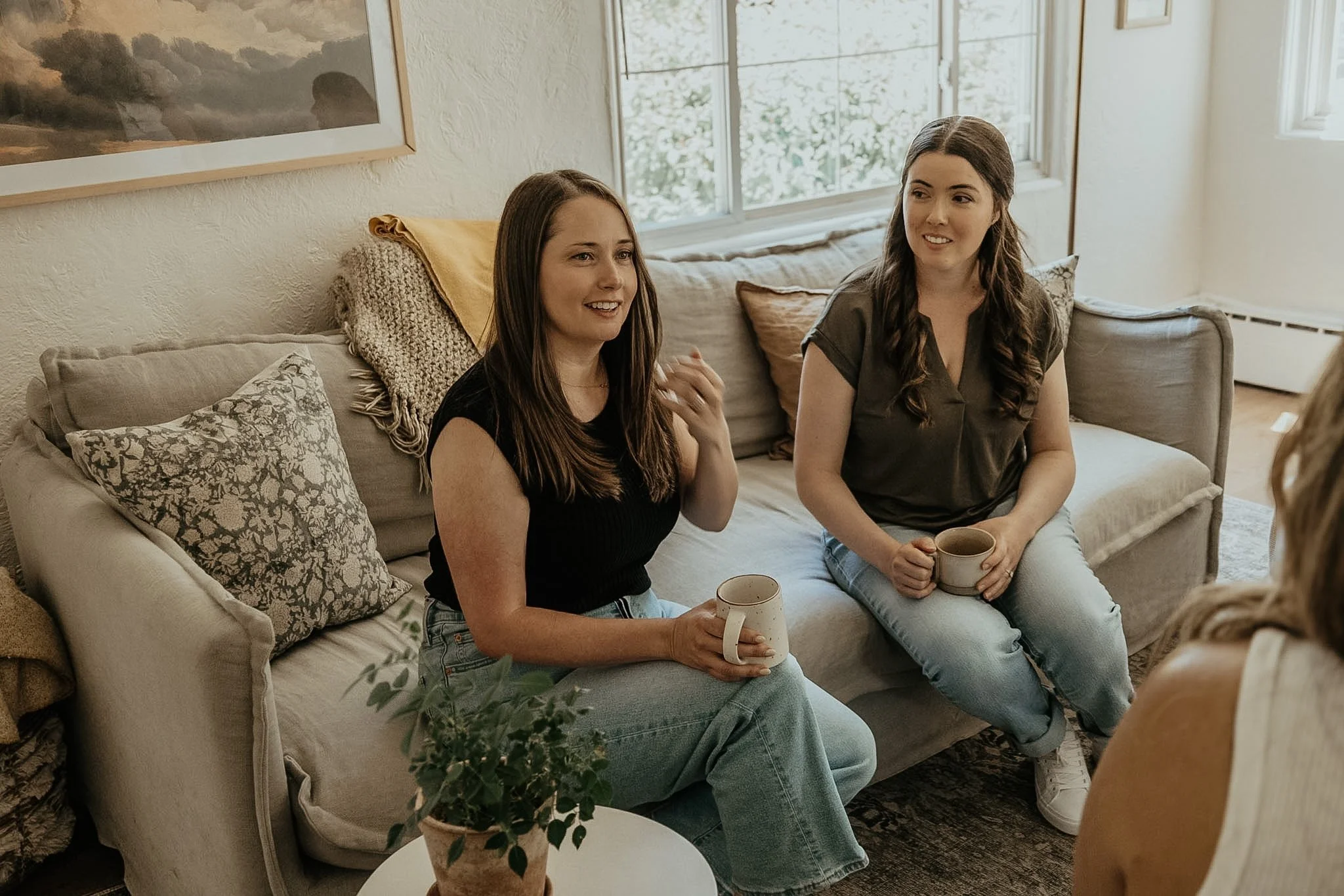 Two women sitting on a beige sofa with patterned pillows, holding mugs, engaged in conversation in a sunlit living room with a large window and framed artwork on the wall.
