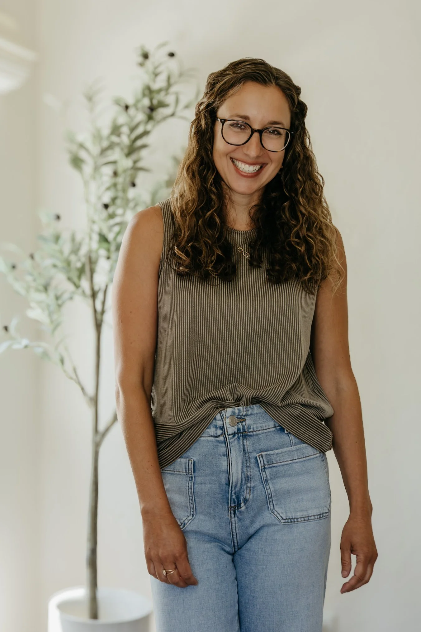 A woman with curly brown hair, glasses, and a warm smile, wearing a sleeveless striped top and light blue jeans, standing indoors near a potted plant.