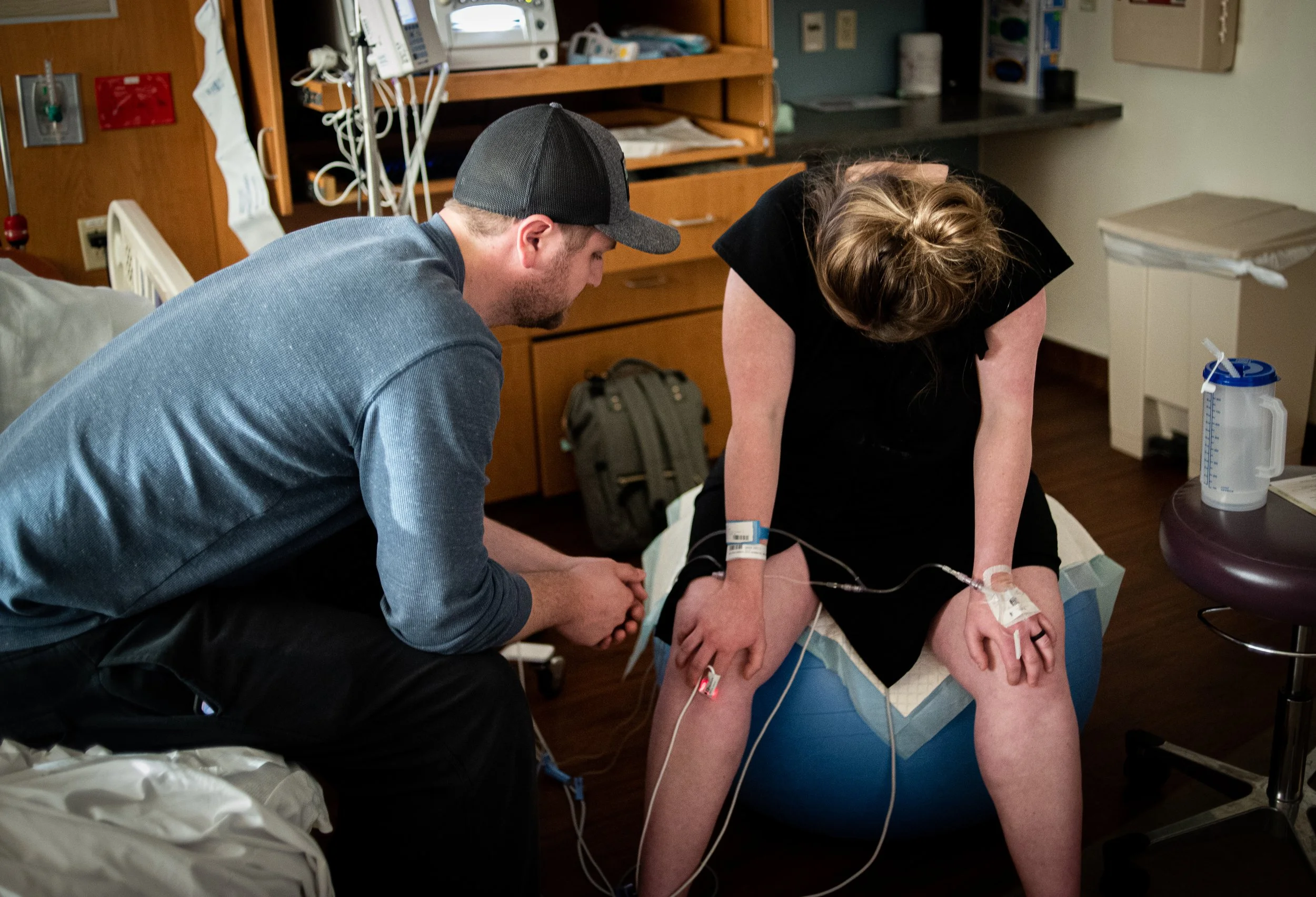 A person is lying on a hospital bed with an IV line in her arm, while a man is sitting and leaning over her, possibly talking to her or checking her condition, in a hospital room with medical equipment and supplies.
