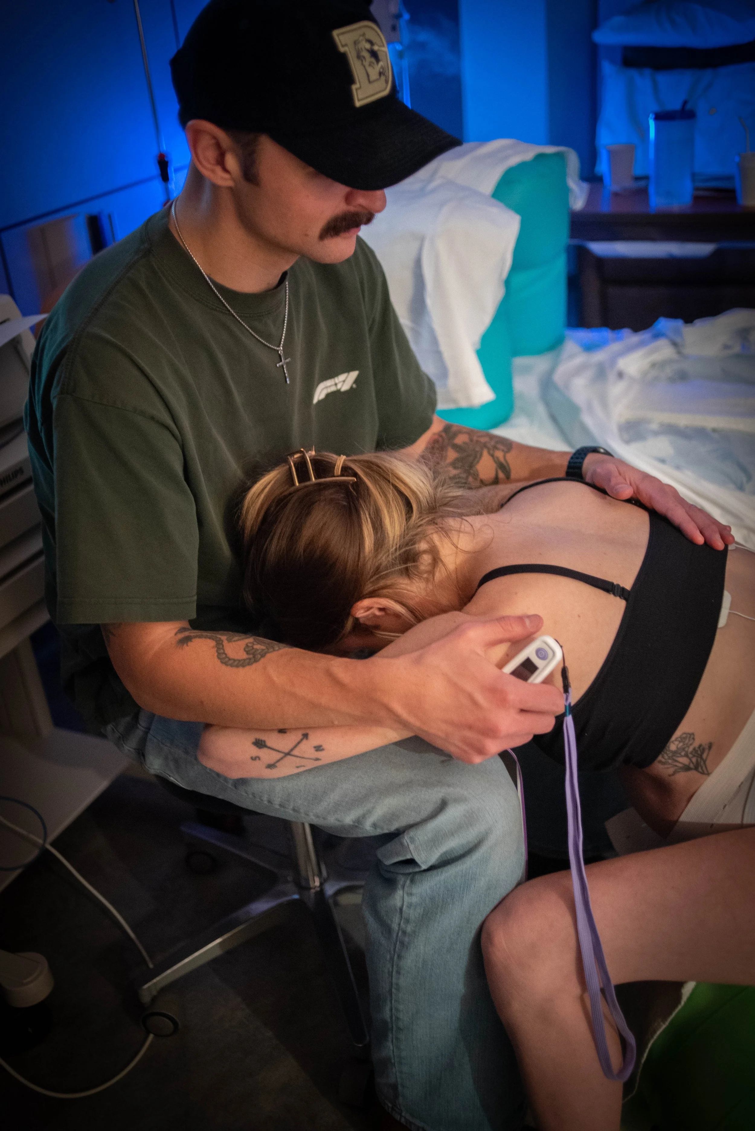 A man with tattoos and a mustache giving chest compressions to a woman lying on her side in a medical setting with IV bags and medical equipment nearby.