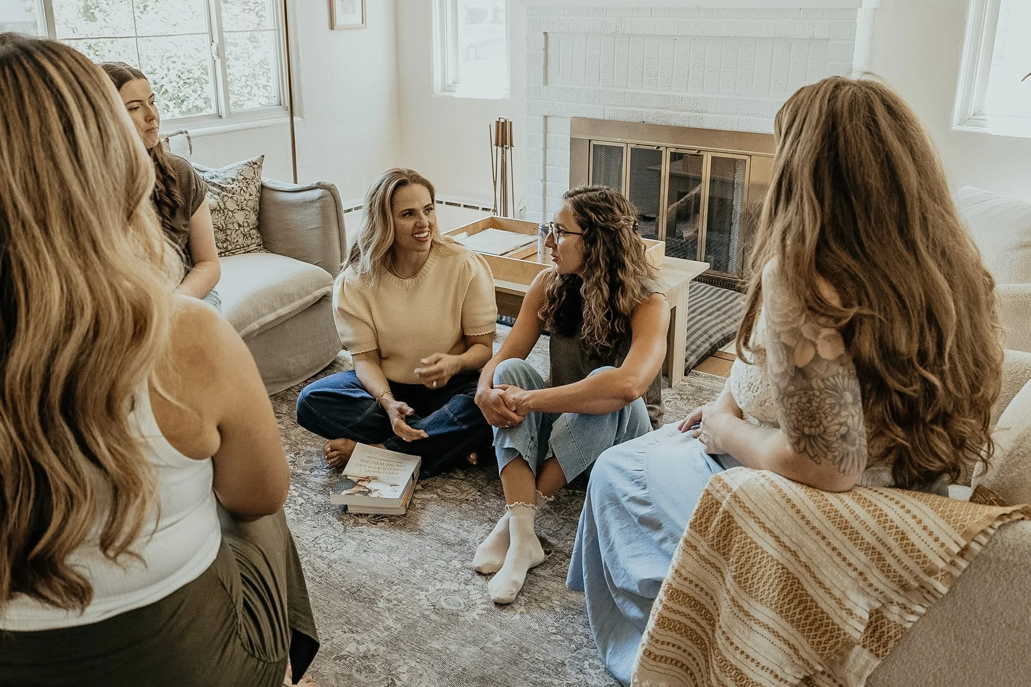 A group of women sitting on the floor in a circle engaging in a discussion in a cozy, well-lit living room.