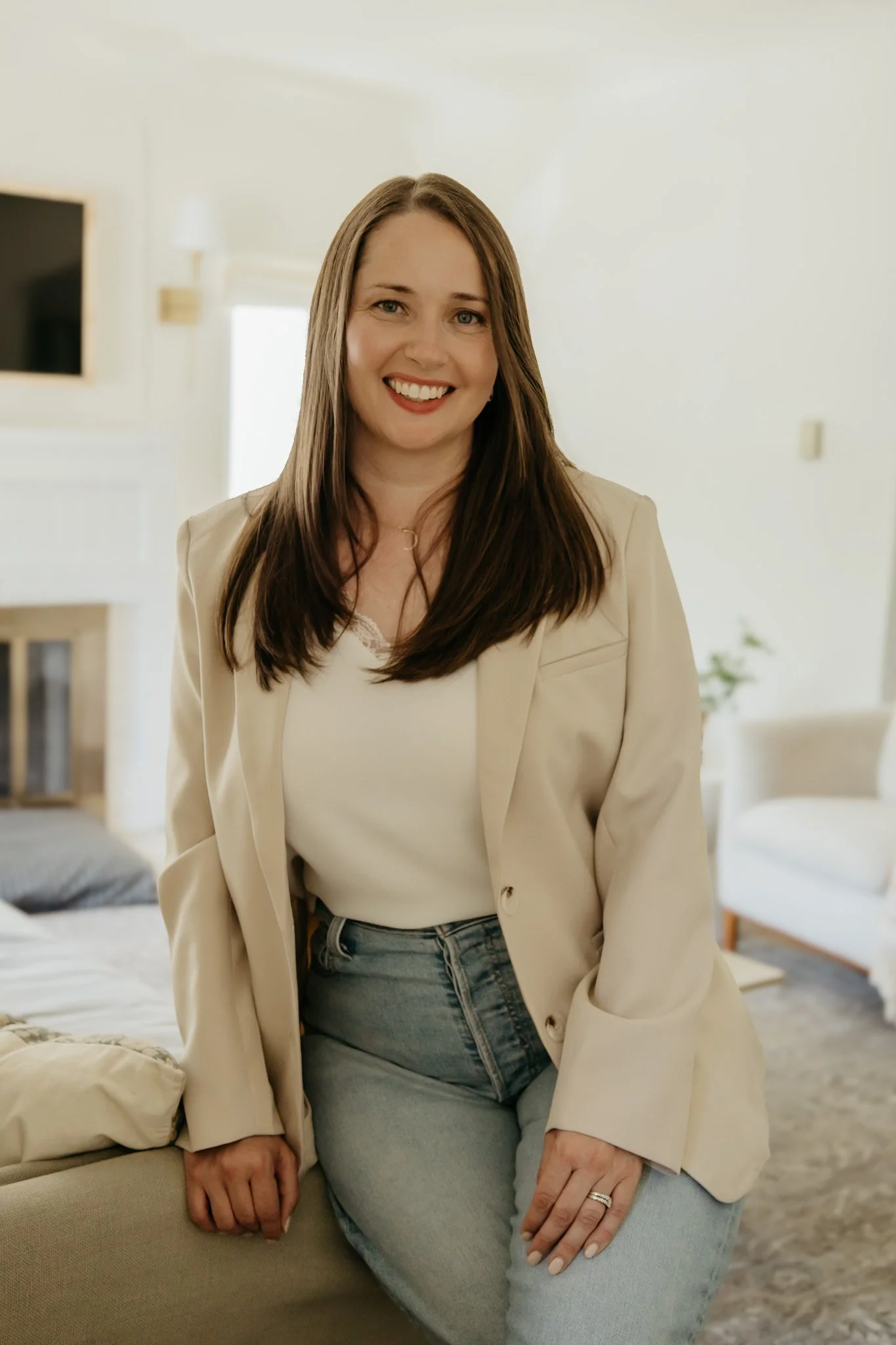 Smiling woman with long brown hair, wearing a beige blazer and light blue jeans, sitting on a beige sofa in a bright, modern living room.