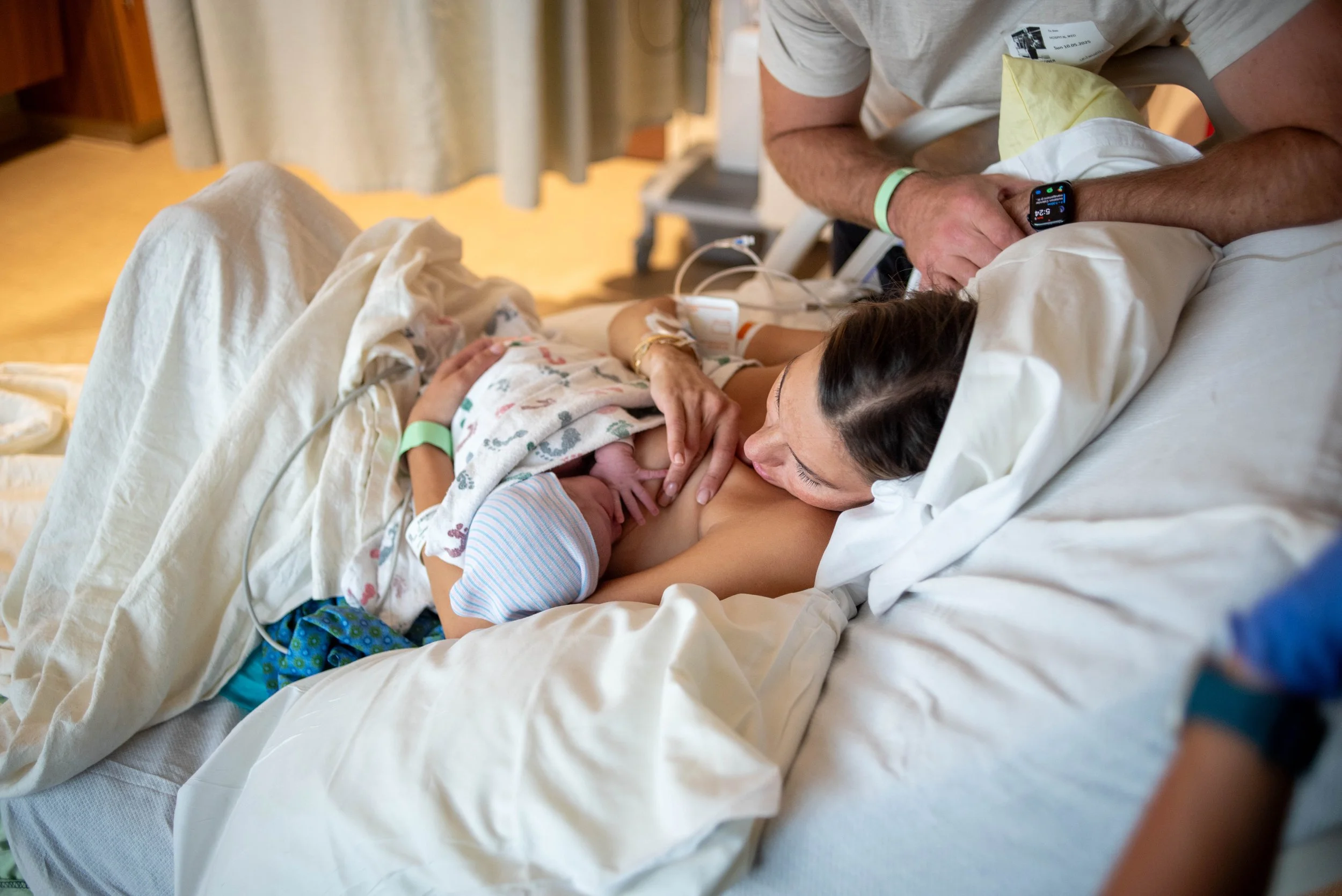 A woman in a hospital bed breastfeeding a newborn baby, with a man beside her holding the baby, wearing a smartwatch and a hospital wristband.