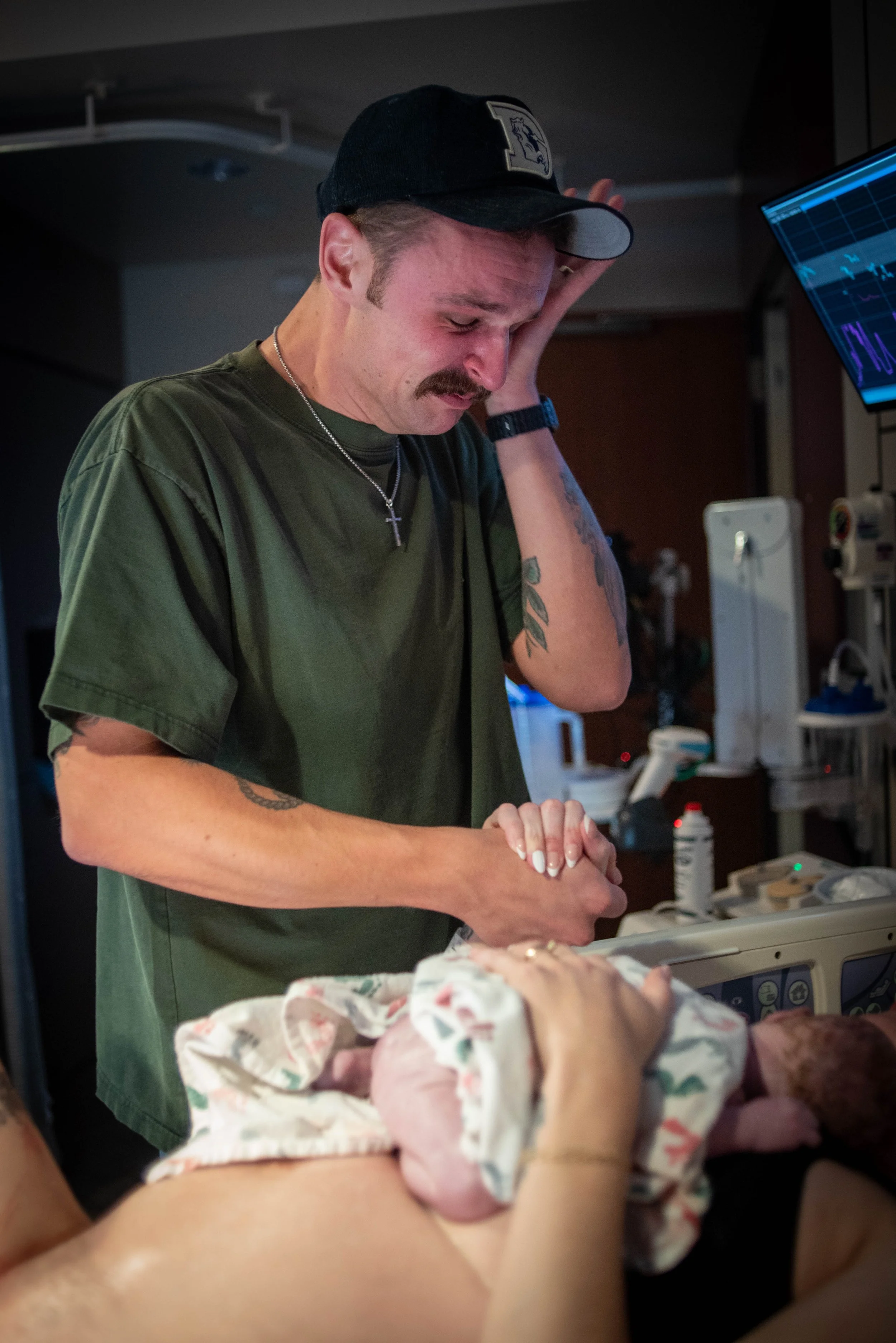 A man wearing a baseball cap, green shirt, and cross necklace, looking down with a distressed expression, is in a hospital room next to a newborn baby wrapped in a pink blanket with a floral pattern, and medical equipment in the background.