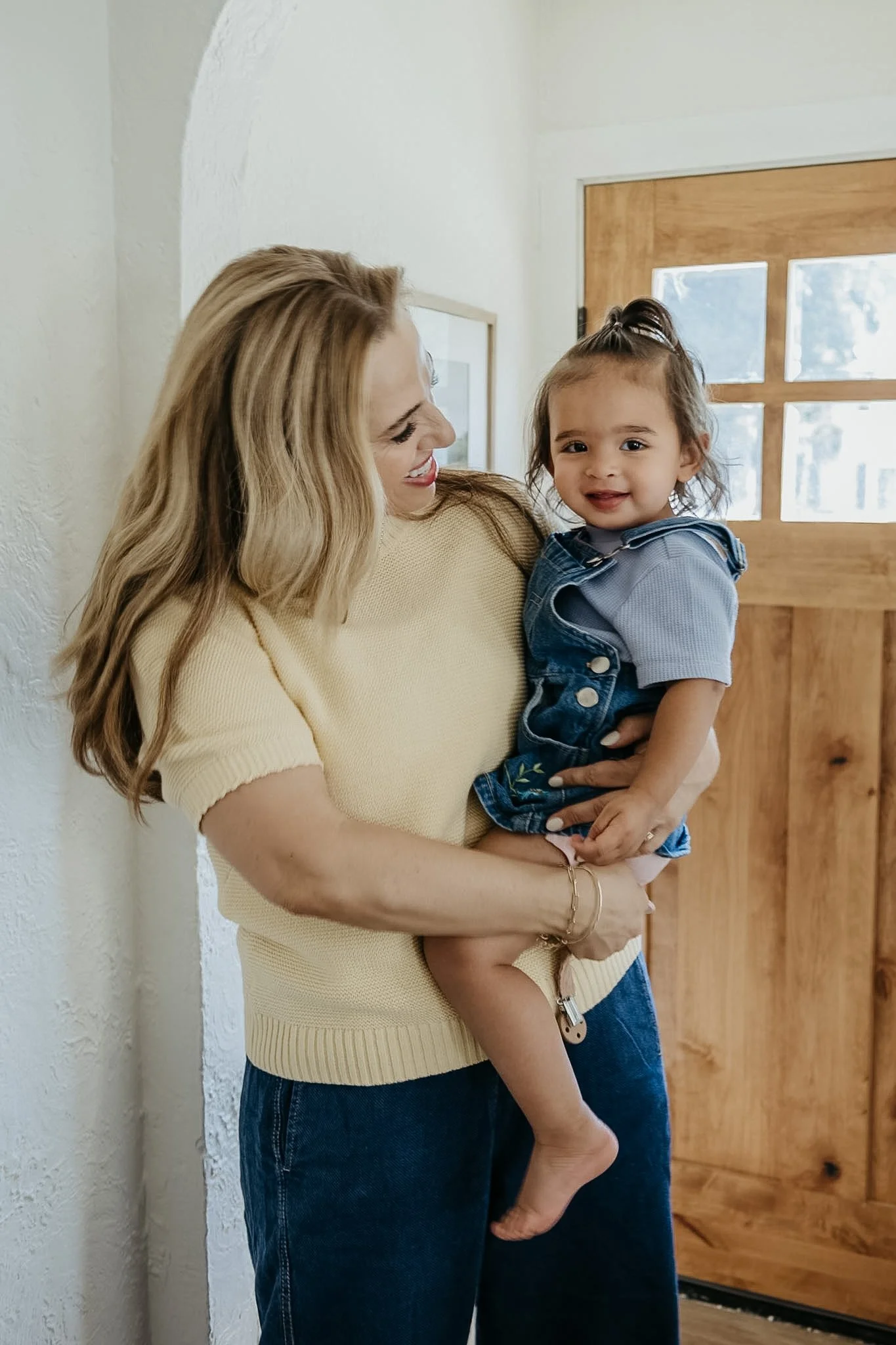 A woman with long blonde hair smiling as she holds a young girl with dark hair, who is smiling and looking at the camera, in a room with a white textured wall and a wooden door in the background.