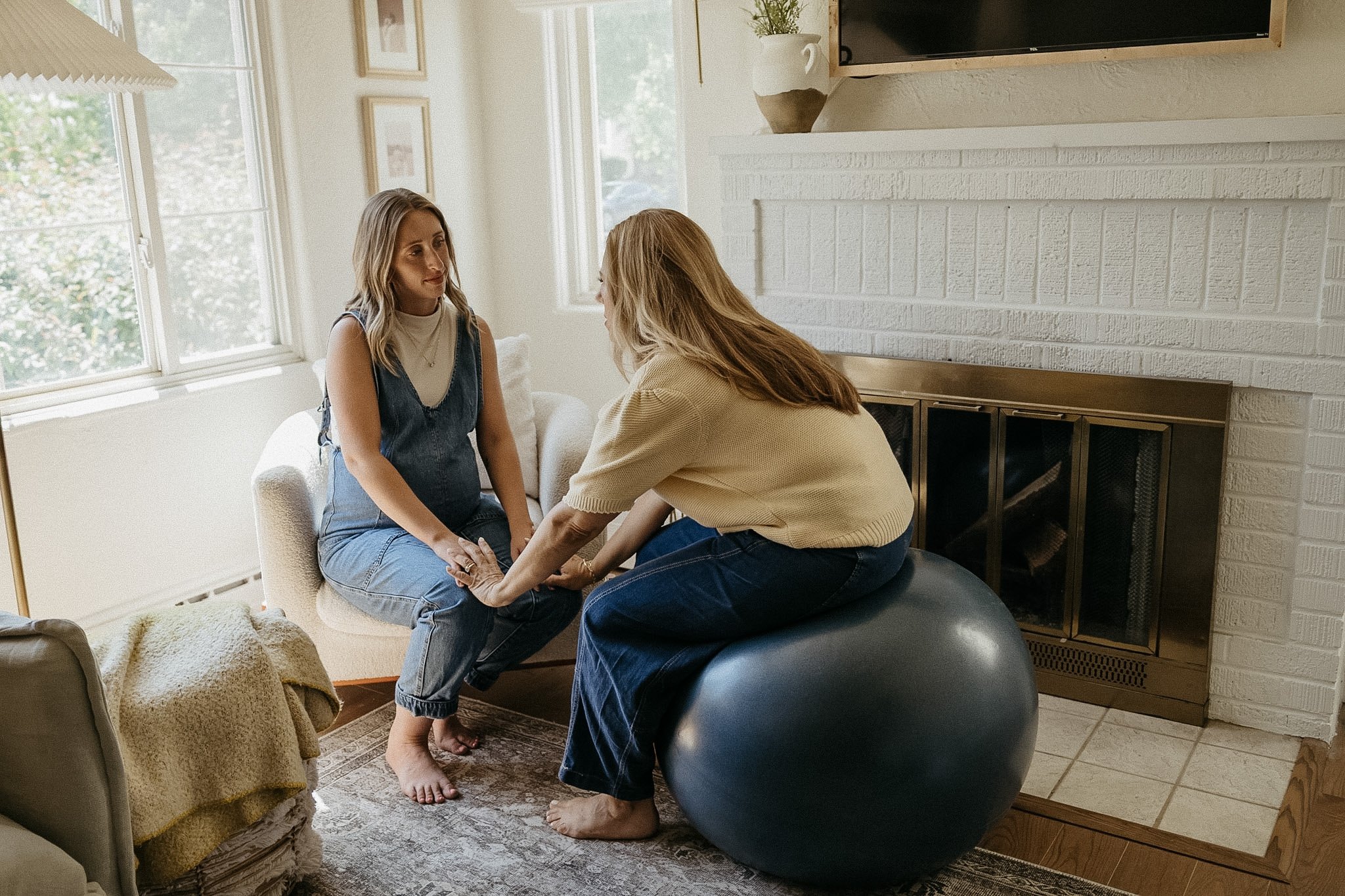 Two women sitting inside a living room, engaging in a serious conversation. One woman is holding the other's hands, and the woman being held appears emotional. The room features large windows, a fireplace, and a wall-mounted TV.