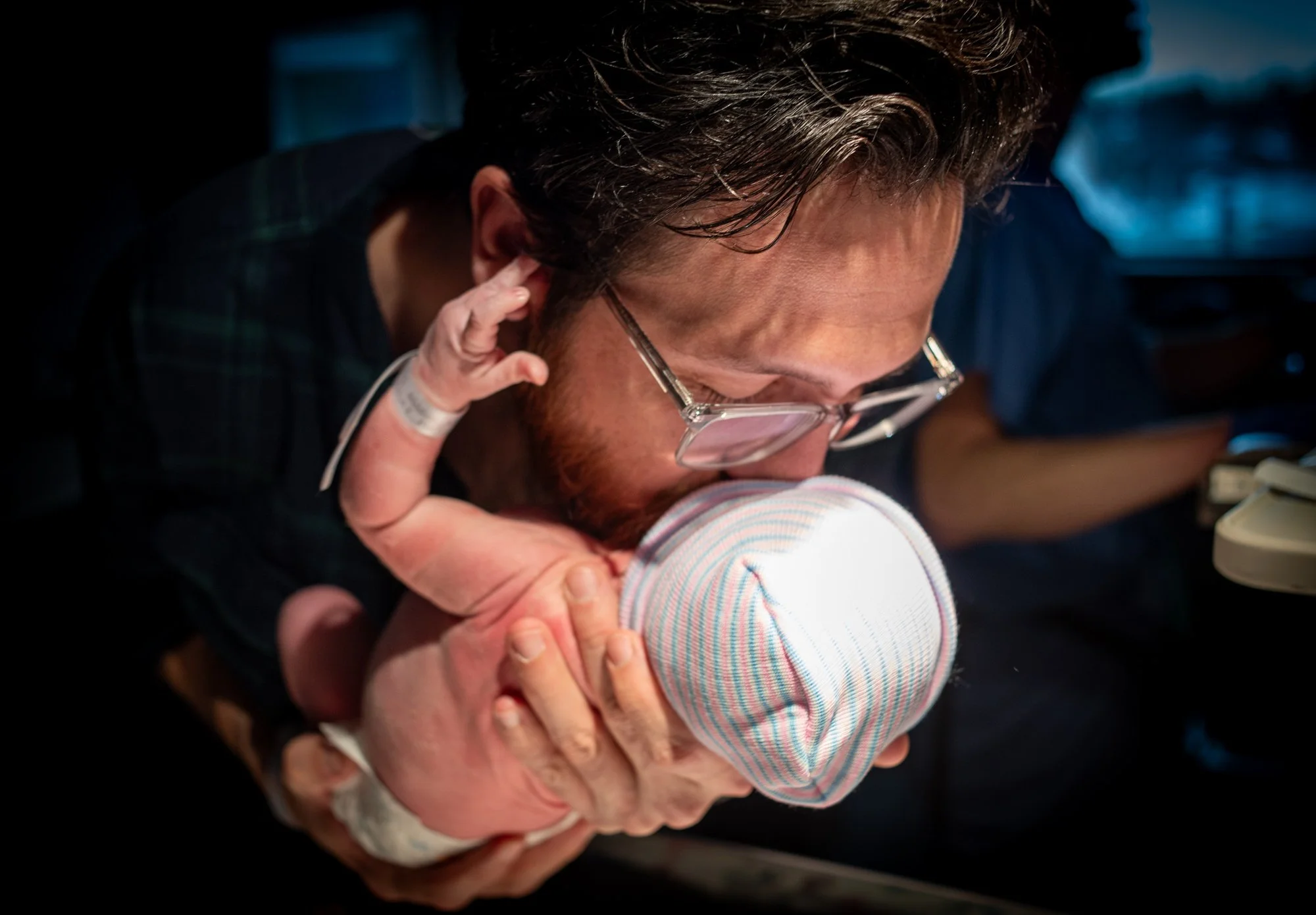 A man wearing glasses is holding a newborn baby dressed in a striped outfit, and the man is kissing the baby's head.