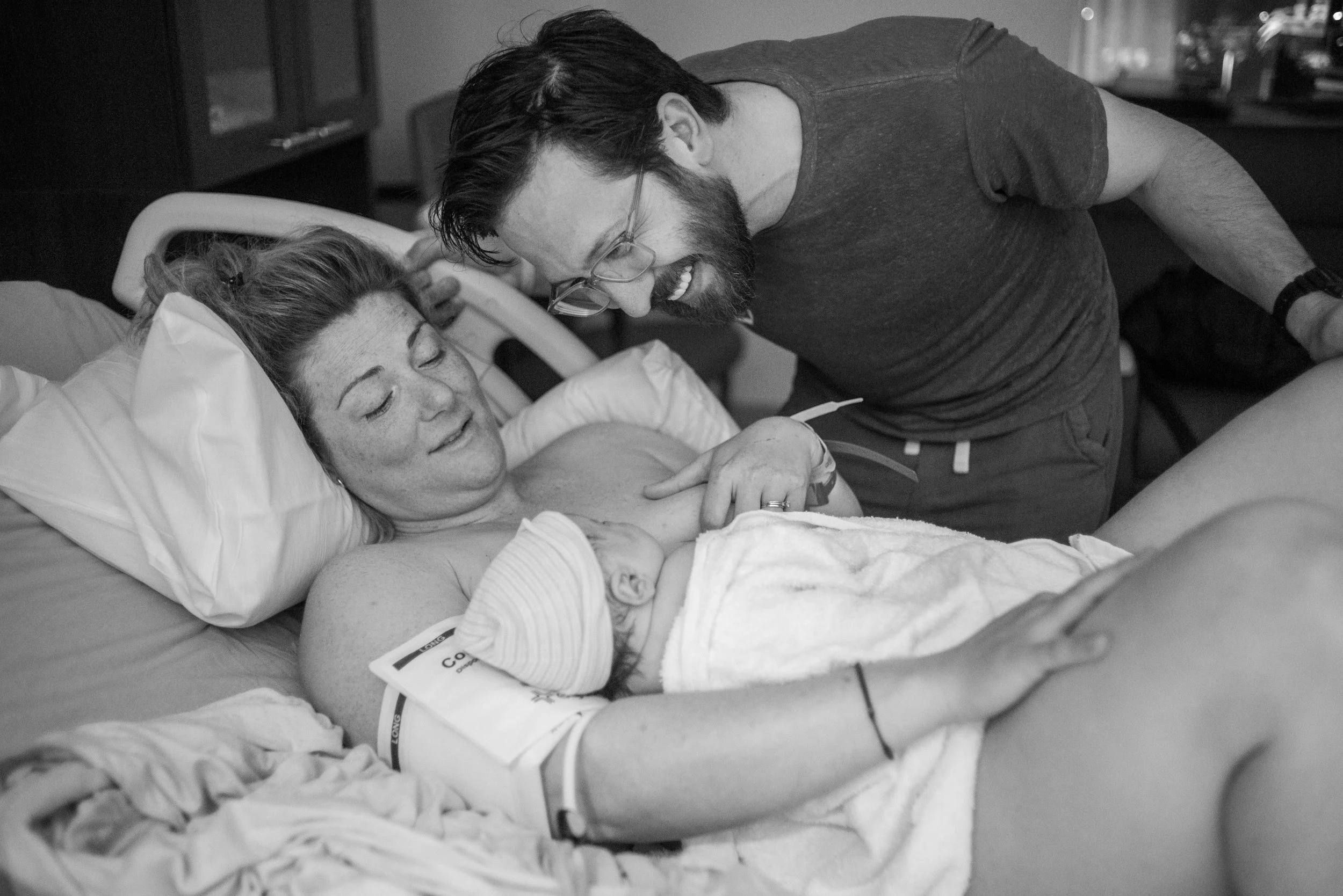 A woman lying in a hospital bed, smiling, with a man leaning over her, appearing to talk or laugh. The scene is in black and white.
