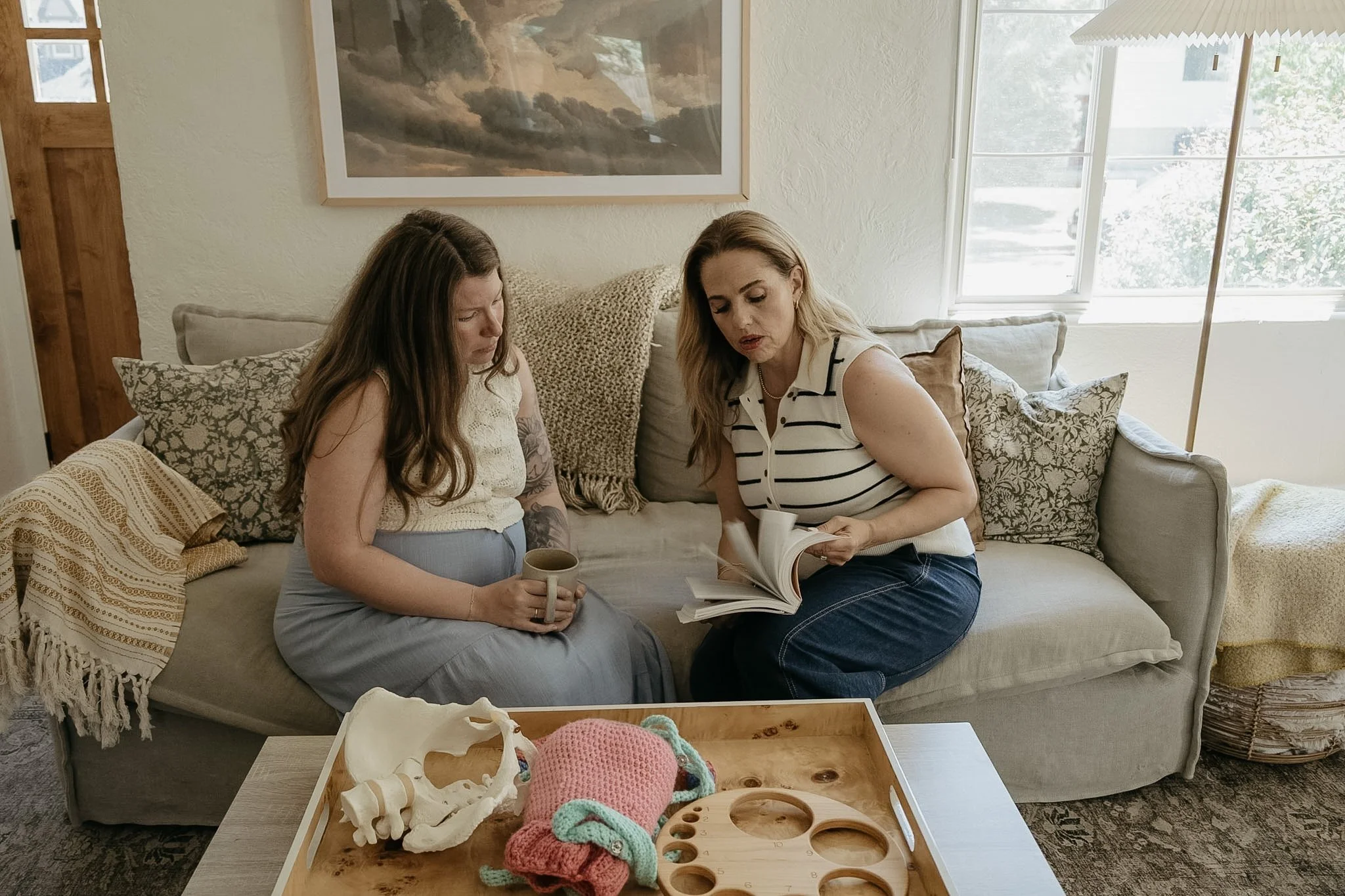 Two women sit on a beige couch in a living room, looking at a book. One woman holds a coffee mug, and the other points at the book. A wooden tray with plastic animal bones and a knitted pink and blue toy is on a table in front of them.
