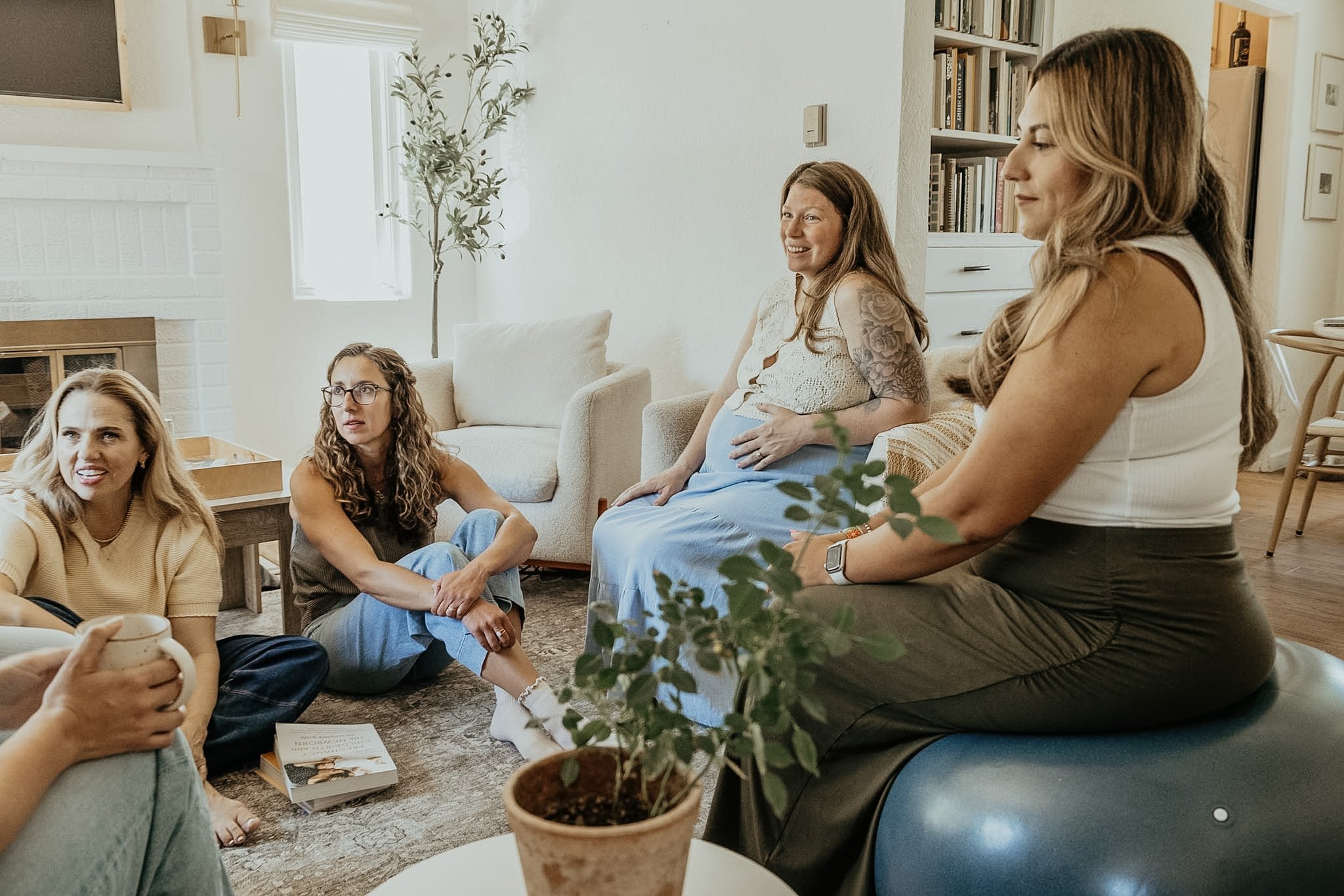 A group of women gathered in a cozy living room, some sitting on the floor, others on sofas, engaging in conversation. A potted plant is in the foreground on a small table, and background elements include bookshelves, a window, and a white sofa.