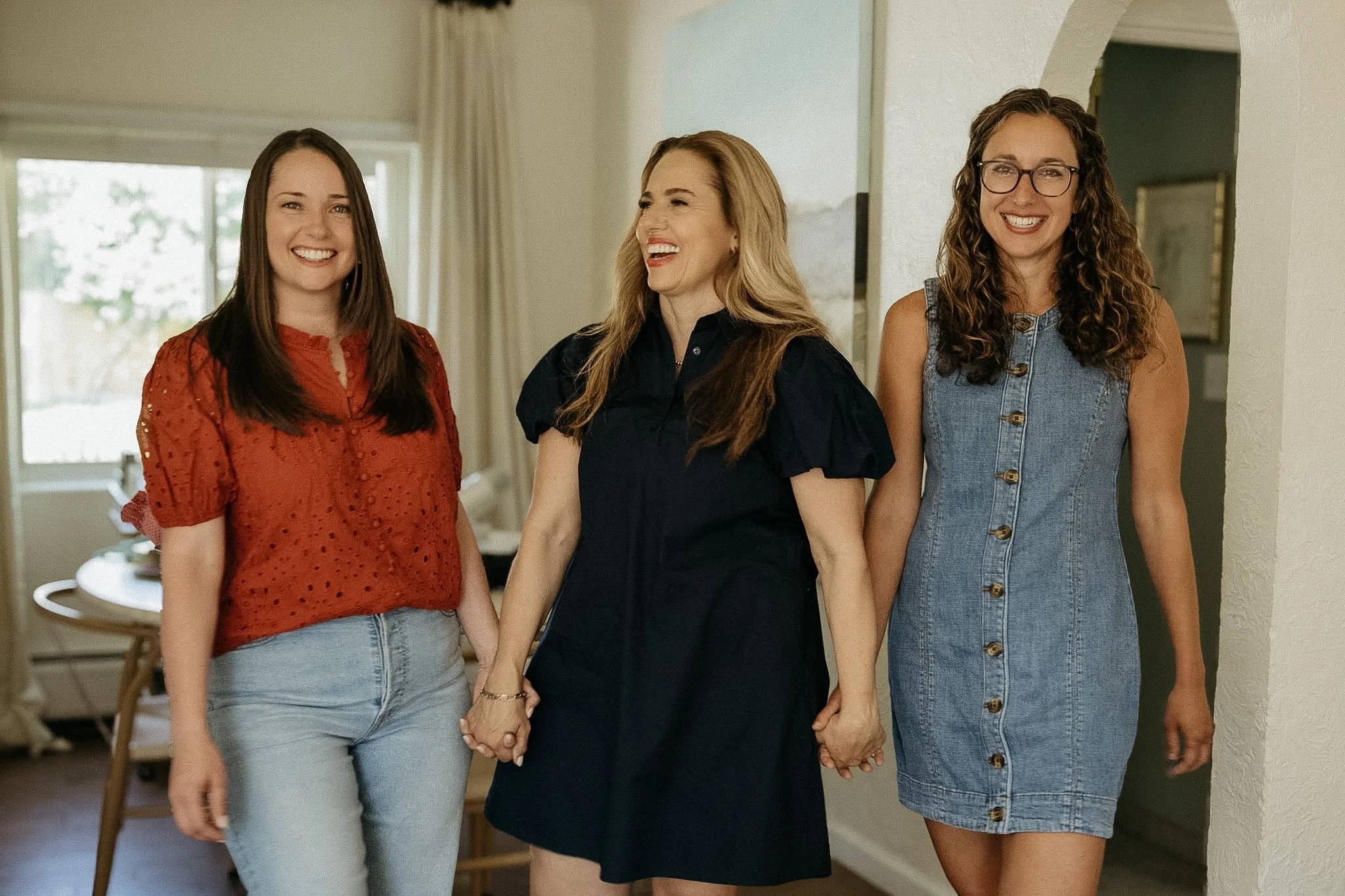 Three women holding hands and smiling in a home, with one woman wearing glasses and a denim dress, another in a black top, and the third in a red blouse and light jeans.