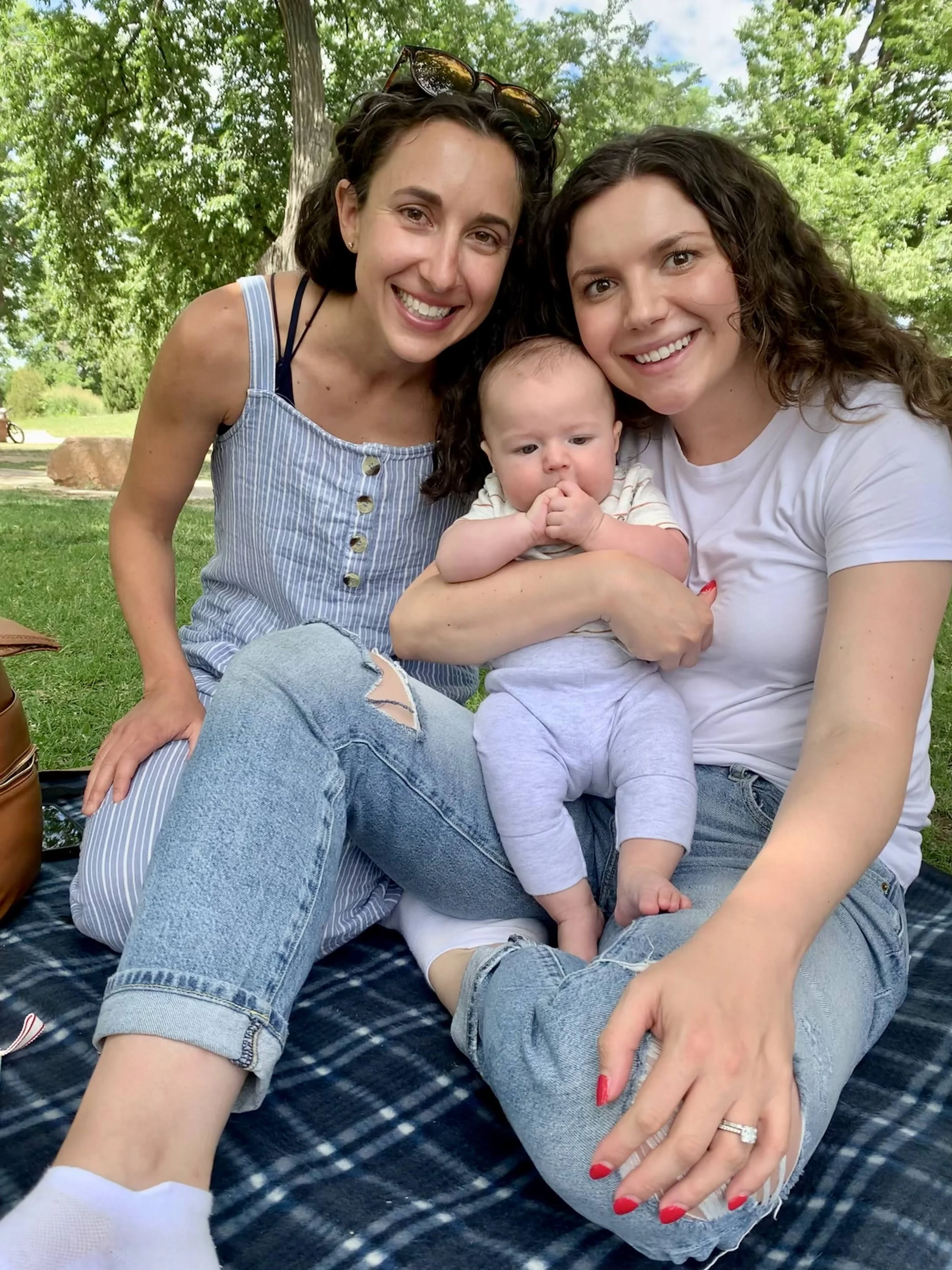 Two women and a baby sitting outdoors on grass beneath trees, smiling at the camera. One woman has curly brown hair and is wearing a white t-shirt, the other has dark hair with sunglasses on her head and is wearing a striped dress. The baby has light hair and is dressed in a striped shirt and gray pants.