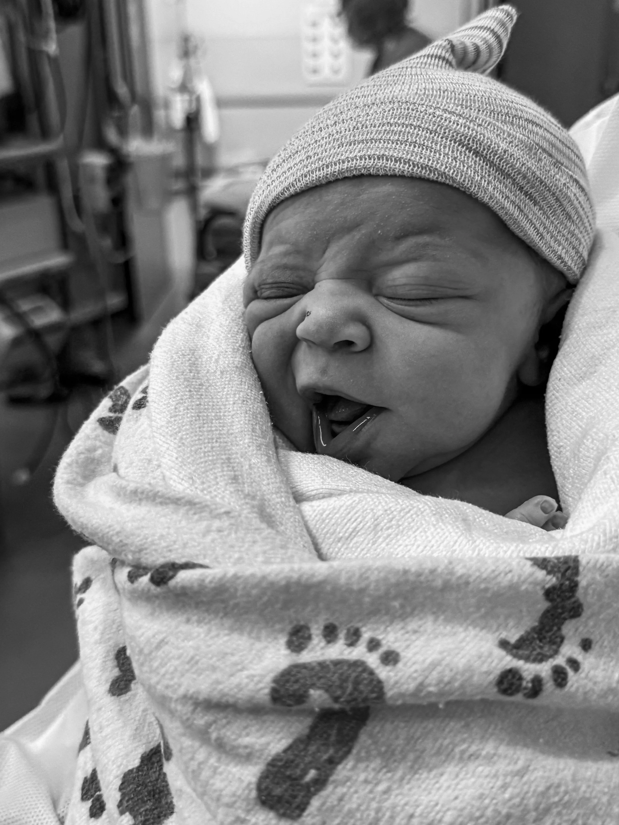 Close-up of a newborn baby’s face with eyes closed, wearing a knit cap, wrapped in a blanket with animal print, lying in a hospital setting.