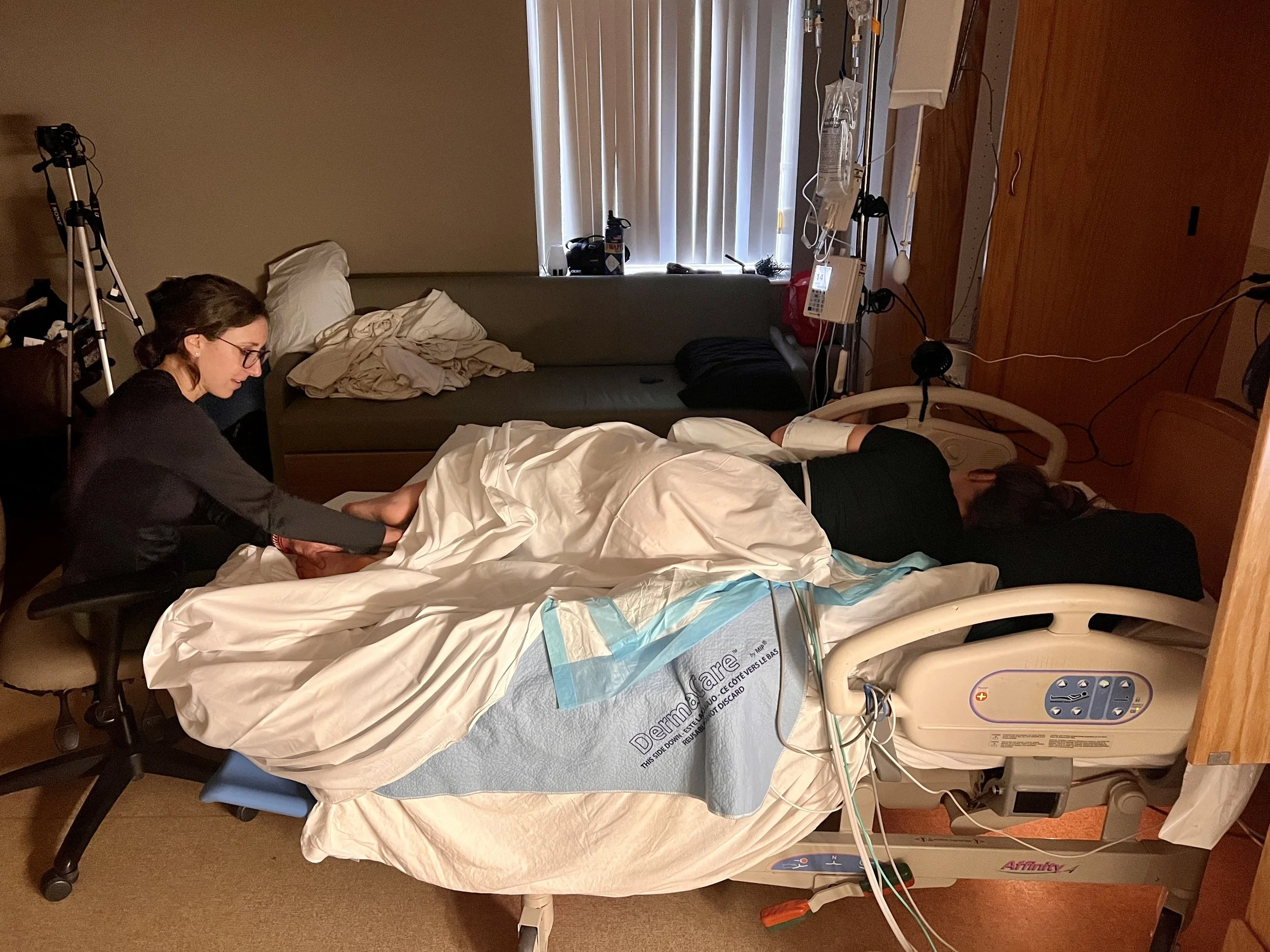 Medical hospital room with a patient on the bed, a woman attending to the patient, and medical equipment including a hospital bed, IV lines, and diagnostic devices.