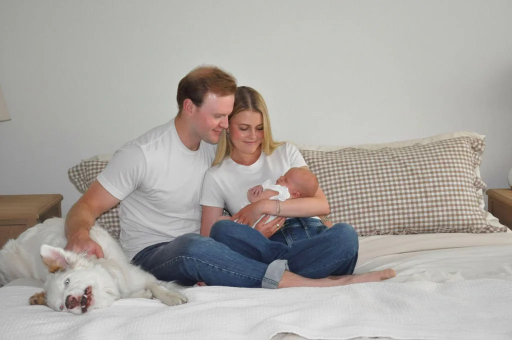 A family of three sitting on a bed with their sleeping dog. The woman is holding a newborn baby.