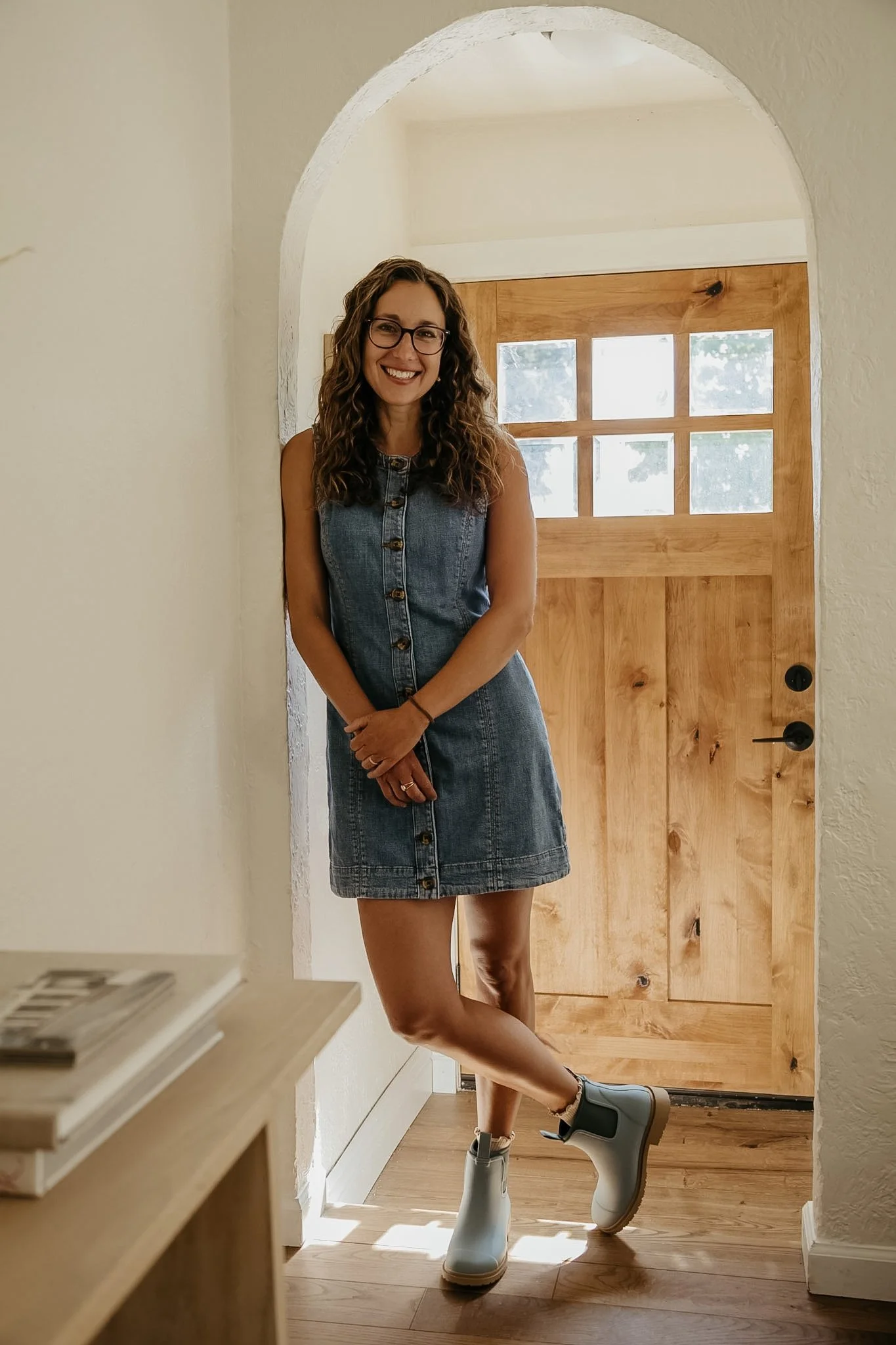 A woman with curly hair, glasses, wearing a denim dress, standing in front of a wooden door with six small windows, smiling at the camera.