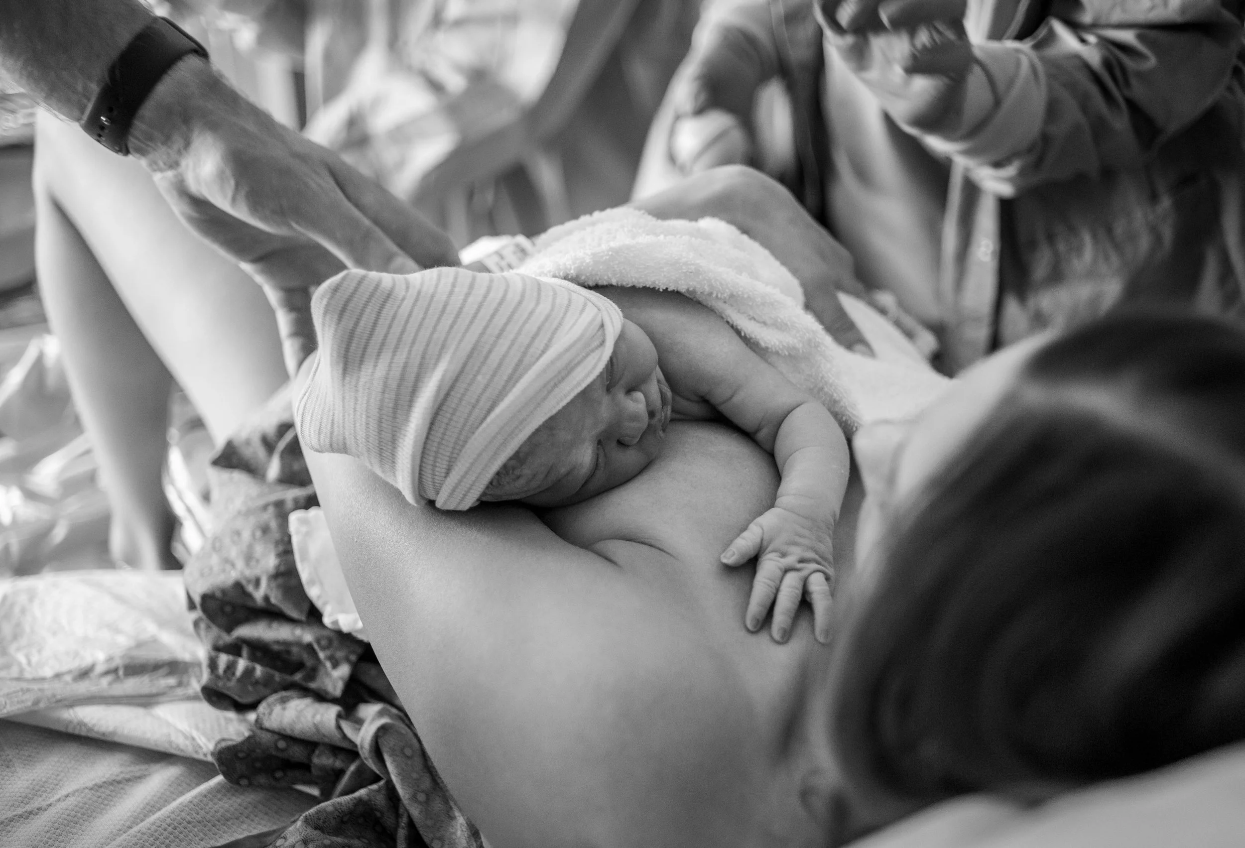 A newborn baby with a striped hat nursing from a person, possibly in a hospital or medical setting, with another person helping or supporting nearby.